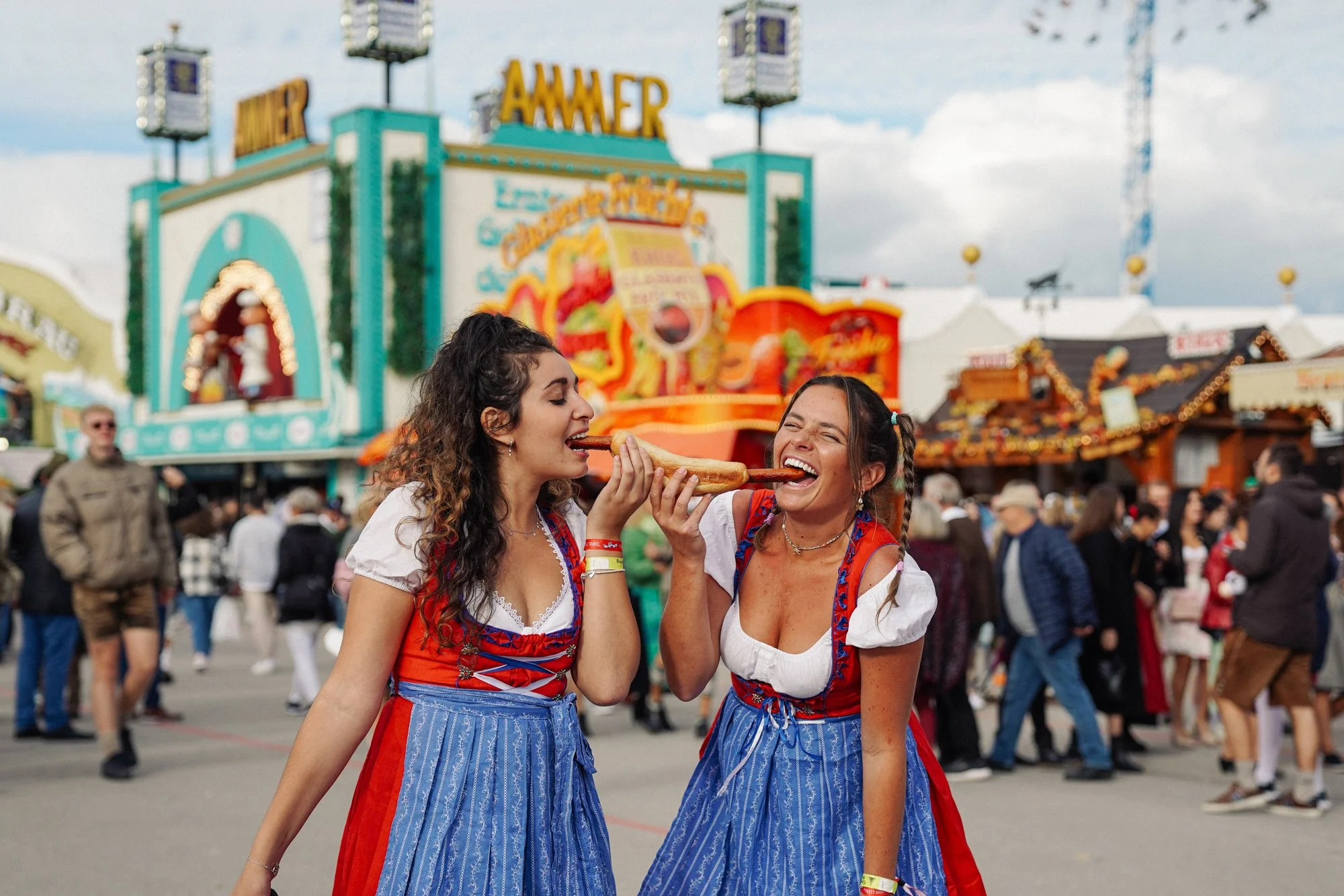 Two women in traditional German dirndls sharing a large pretzel at a carnival or fairground with rides and food stalls in the background.