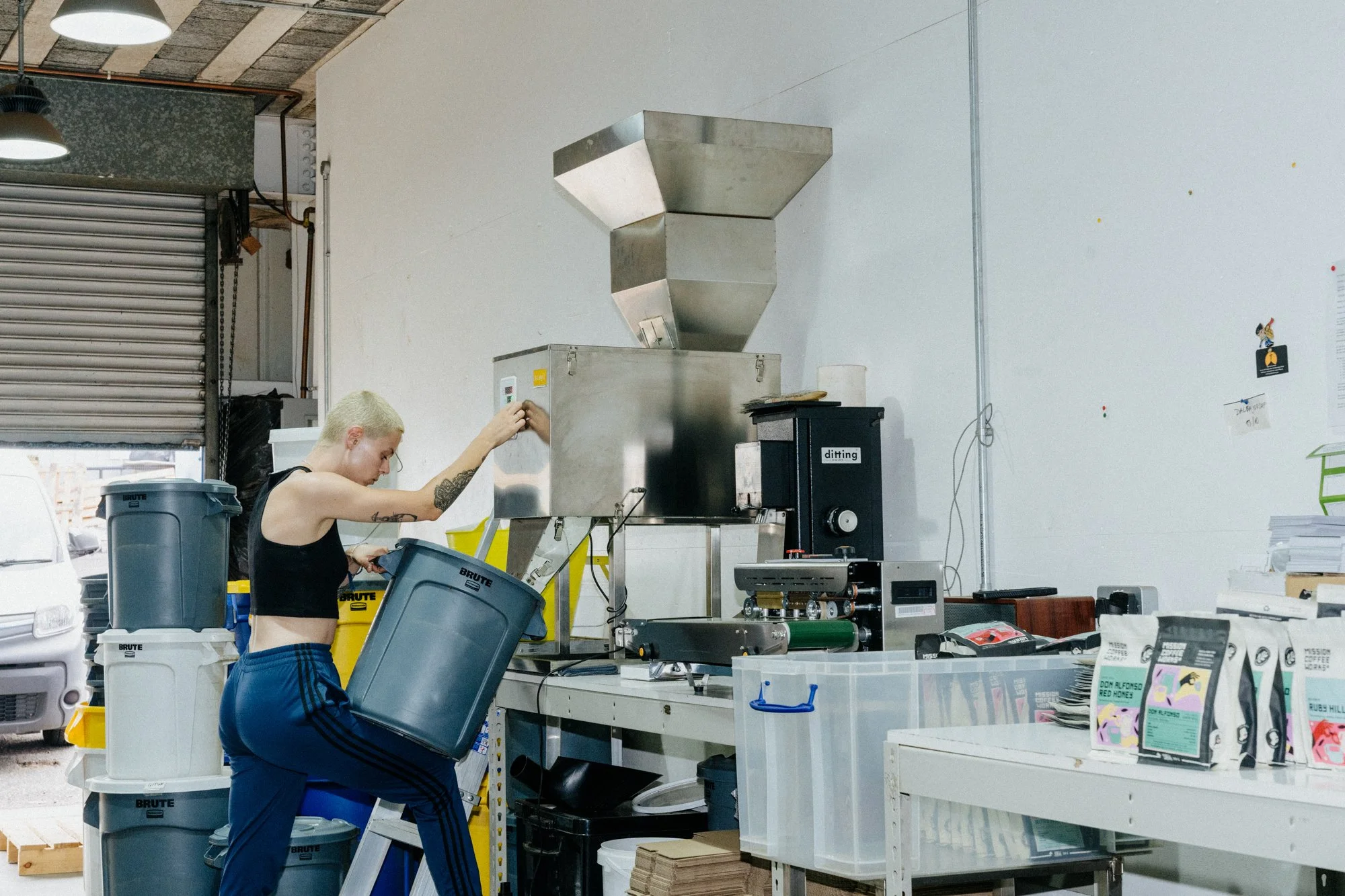 A person with short blond hair and tattoos on their arm, wearing a black crop top and blue striped pants, loading trash into a large gray bin in an industrial workspace. There are various storage containers, machinery, and packages on a white table to the right.