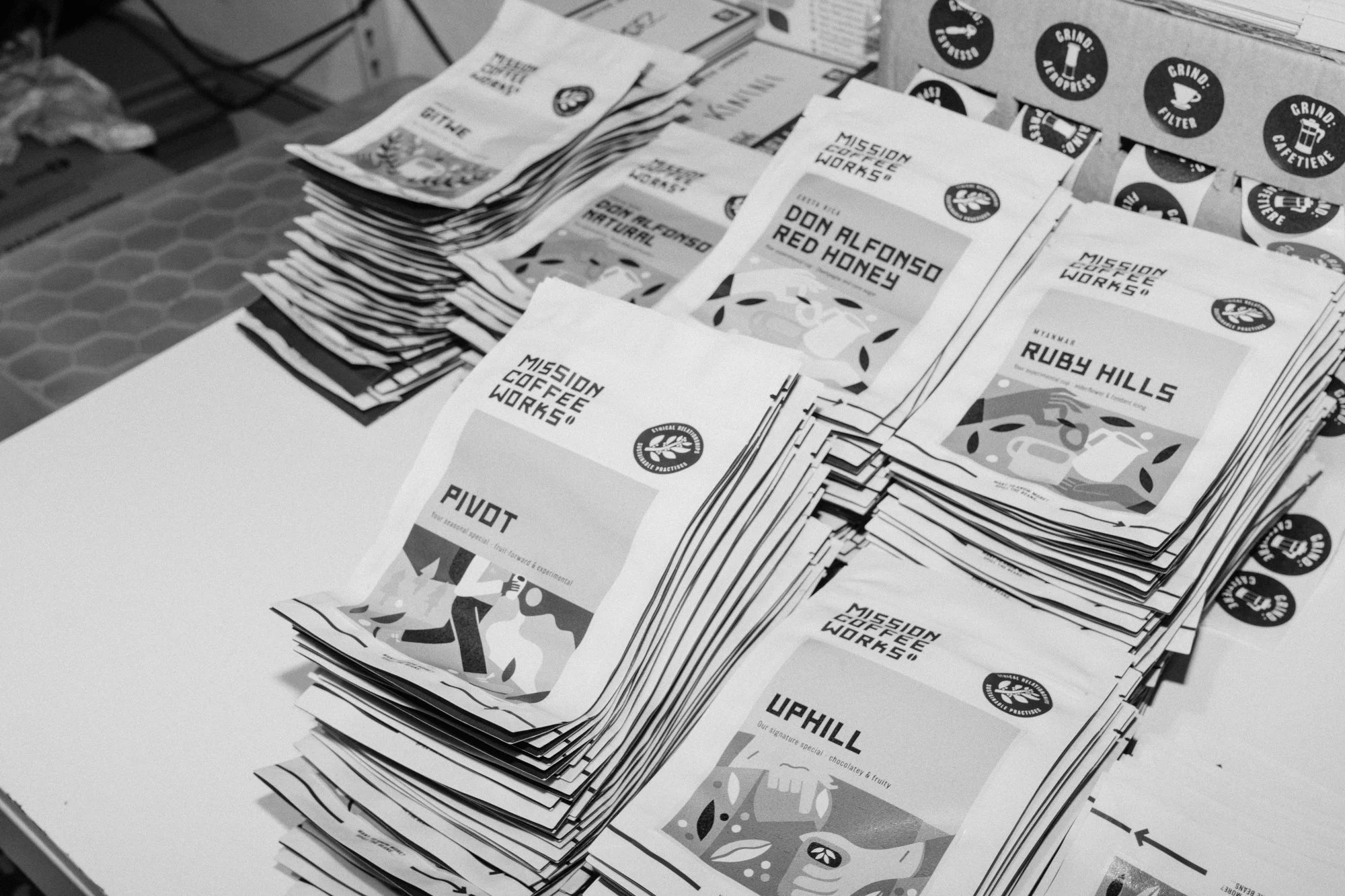 Stacks of packaging paper for coffee blends, including 'Pivot', 'Uphill', and others, with branding for Mission Coffee Works.