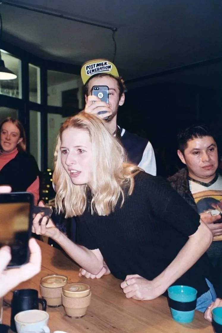 A group of people gathered around a wooden table, with some holding up their smartphones. One woman with blonde hair is leaning forward, looking at her phone. Another man is wearing a yellow button with the words 'Post Milk Generation' on it. The background shows a modern indoor setting with large windows.
