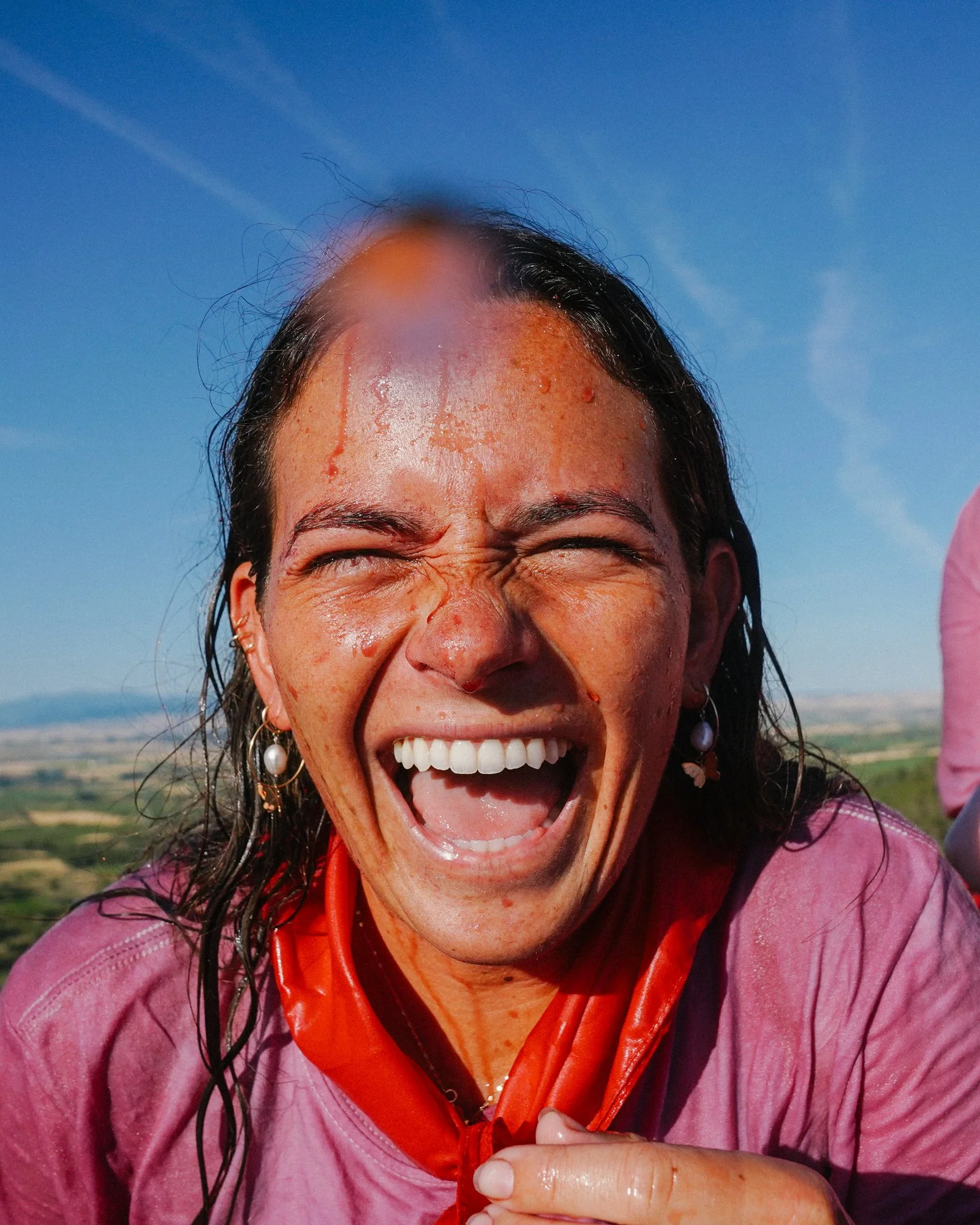 A woman with wet hair and a joyful expression, smiling widely with her eyes closed outdoors on a bright, sunny day.