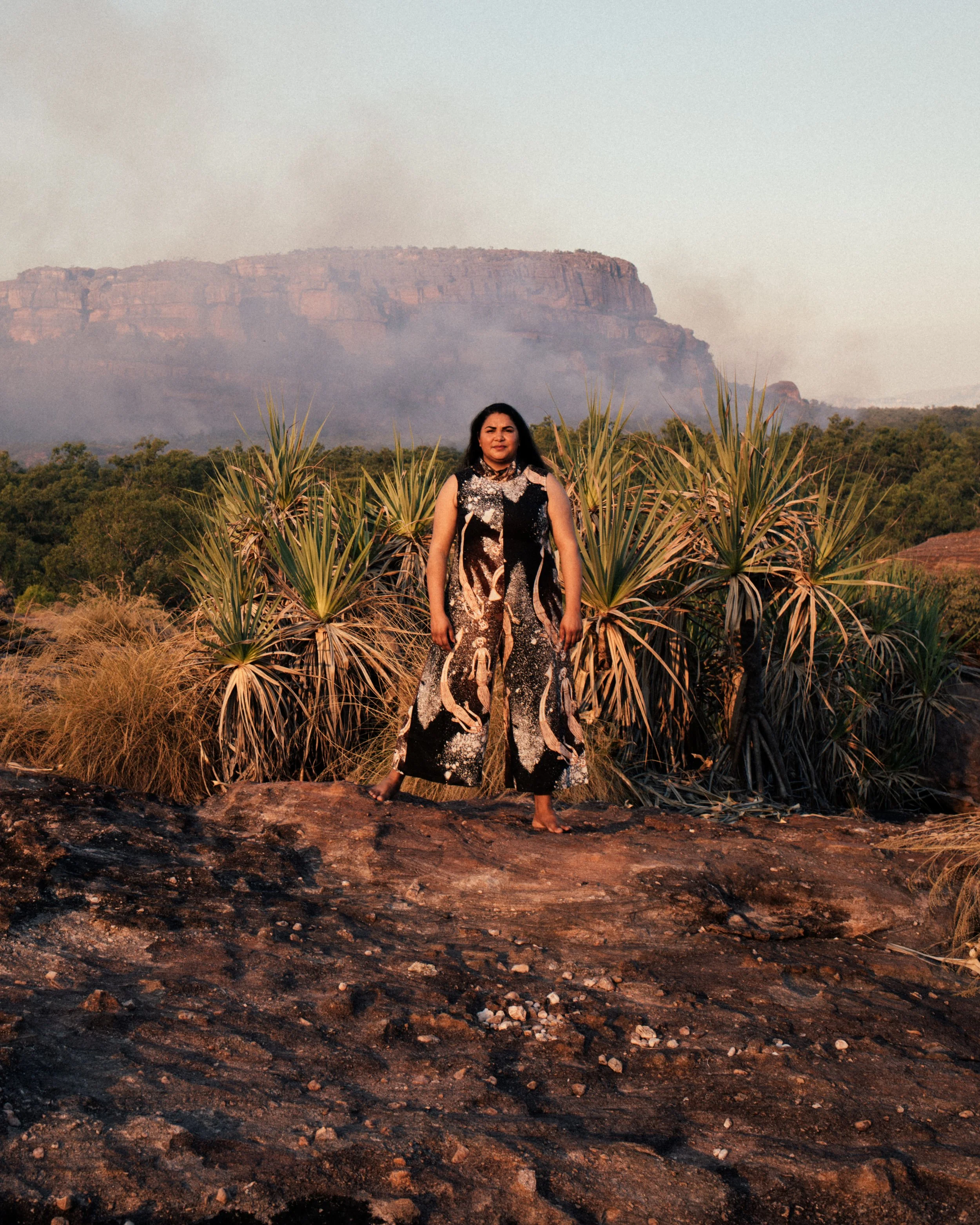 A woman standing on rocky ground in a desert landscape with plants and a large mesa in the background during sunset