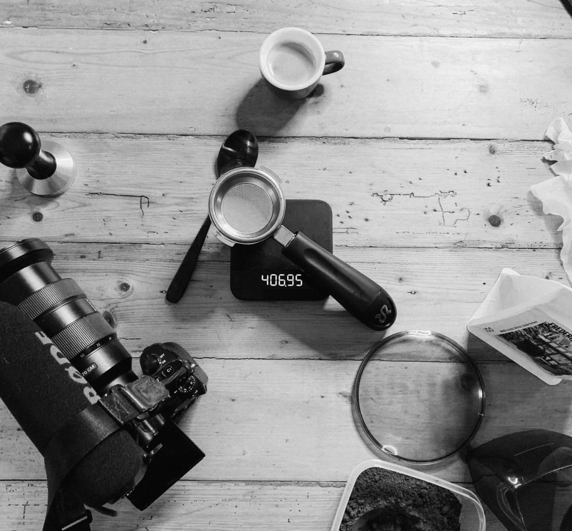 Black and white photo of a wooden table with various objects, including a camera, a coffee tamper, a coffee scoop, a coffee filter, a digital scale showing 406.95 grams, a small cup, a plastic container with ground coffee, a spoon, a paper towel, and a glass bowl.