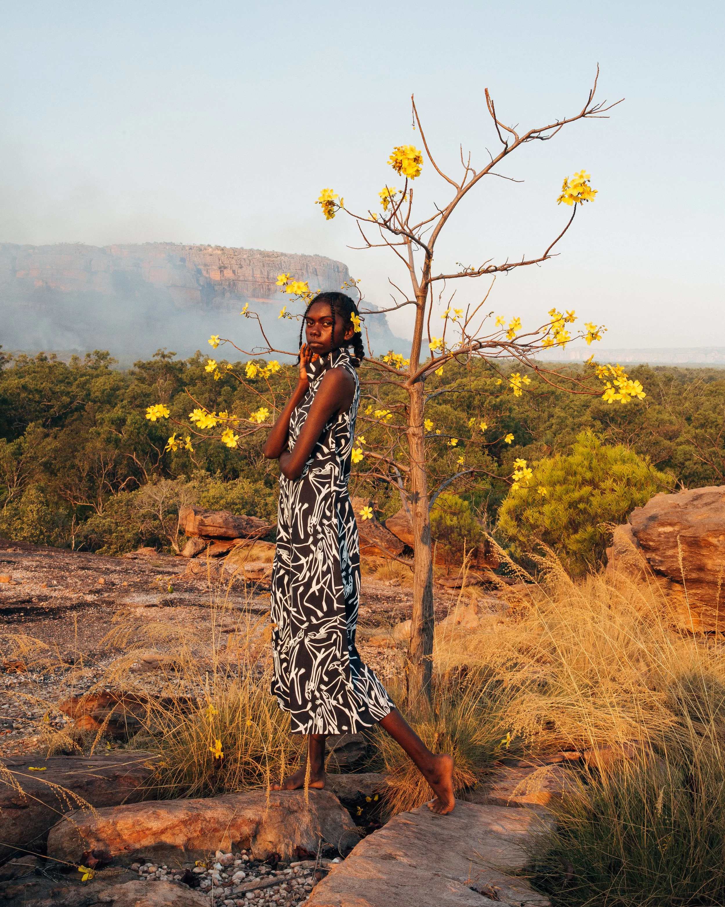 A woman in a black and white patterned dress standing barefoot on rocky terrain near a leafless tree with yellow flowers, with a vast landscape of green trees and distant cliffs in the background during sunset.