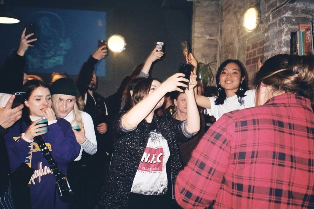 Group of people at a party or social gathering, taking photos and enjoying themselves indoors with brick walls and bookshelves in the background.