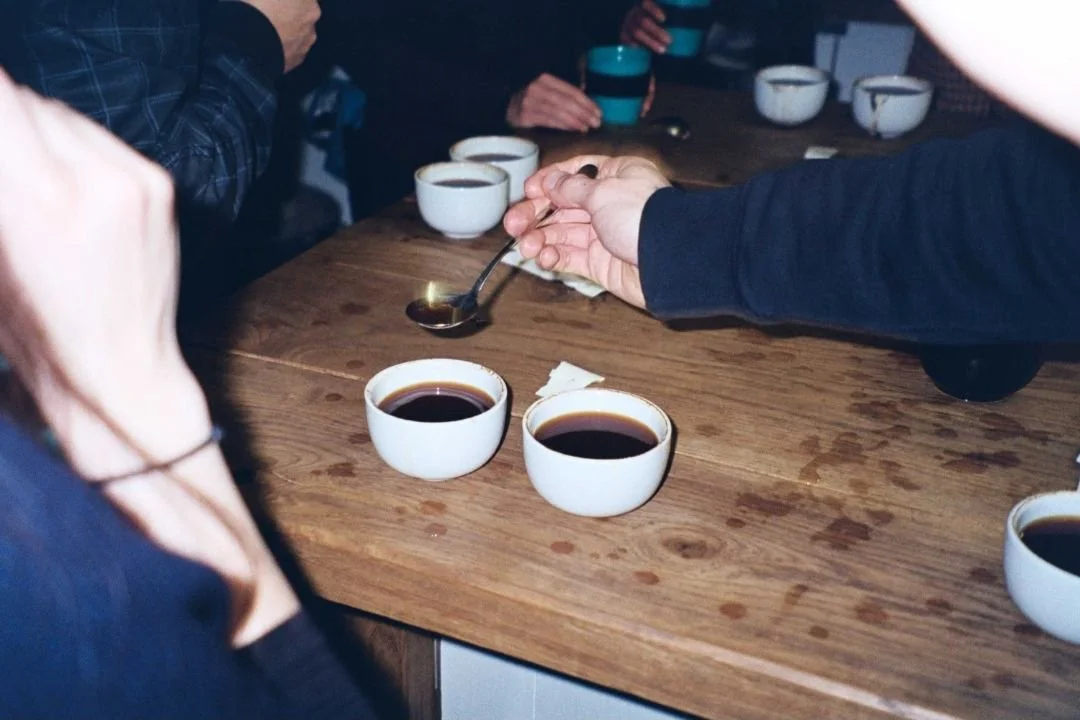 Several people around a wooden table, with cups of black coffee and bowls, one person is scooping coffee from a bowl.