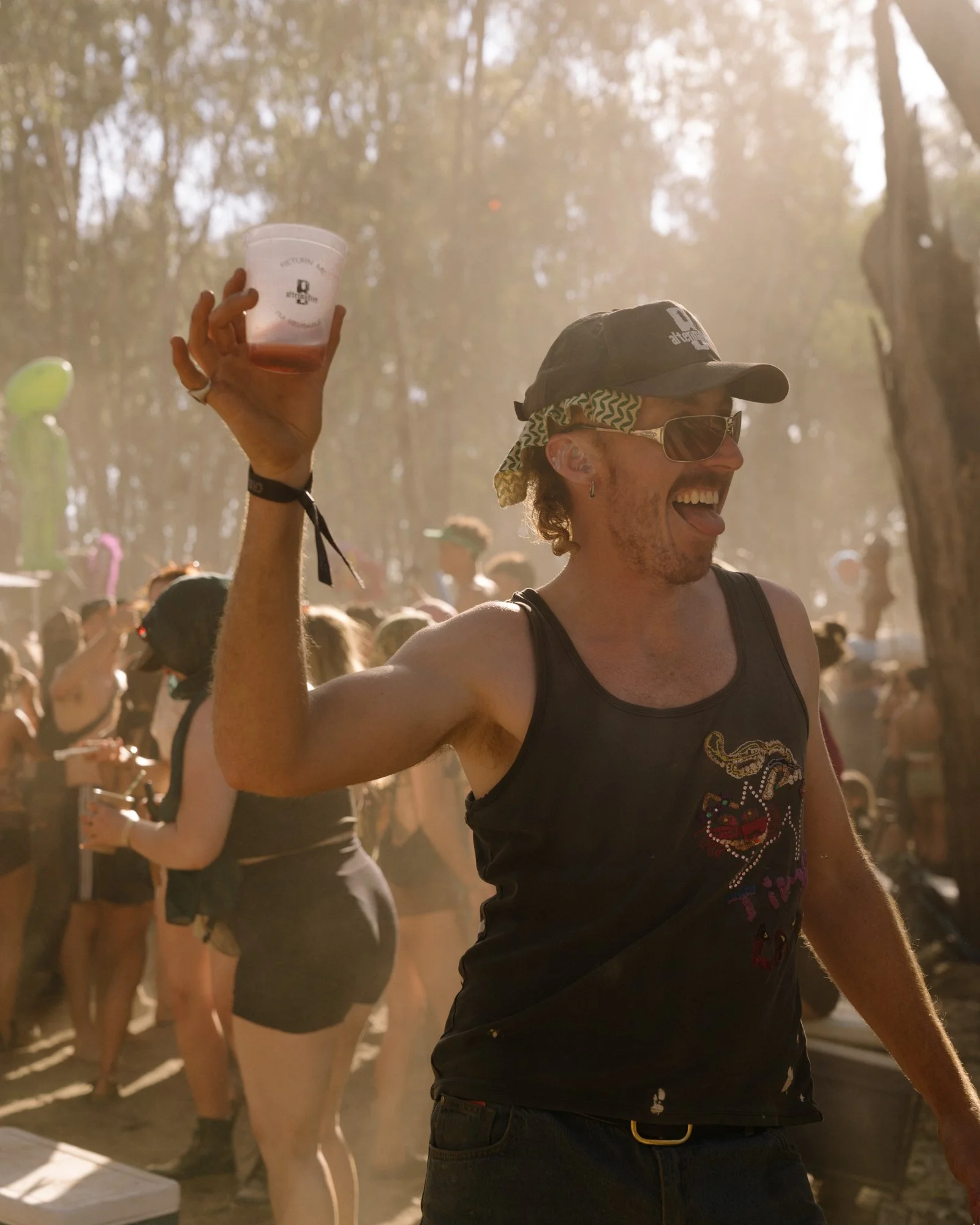 A man wearing sunglasses, a black tank top with a colorful embroidered design, and a black cap, smiling and raising a plastic cup at an outdoor event in a wooded area with a crowd of people around, during daytime.