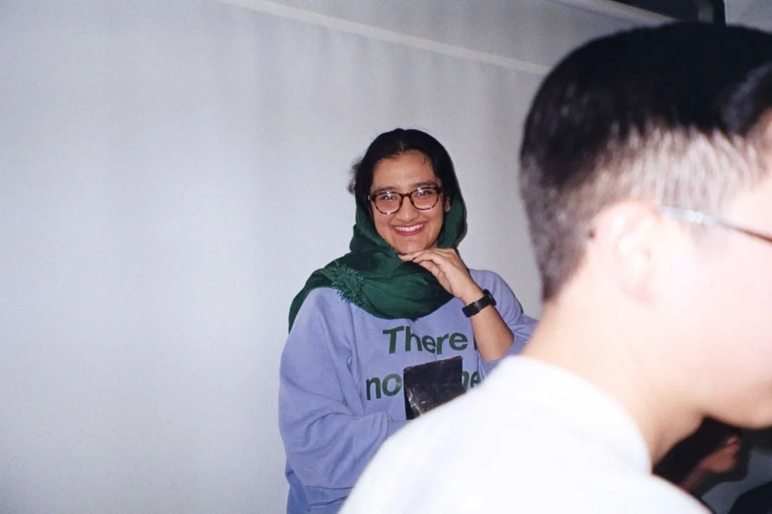Smiling woman wearing glasses and a green headscarf, standing against a plain light-colored wall.