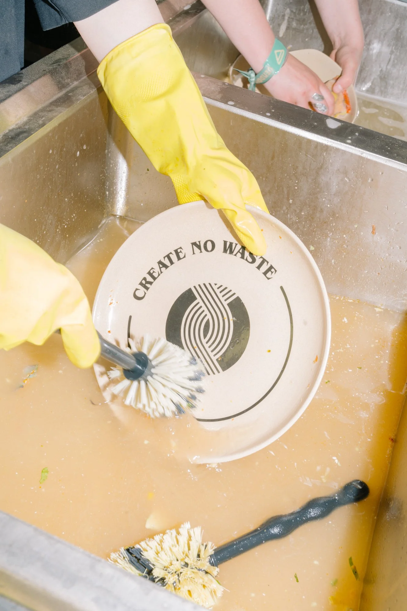 Person wearing yellow rubber gloves washing a plate in a sink filled with soapy water, with a cleaning brush visible in the water.