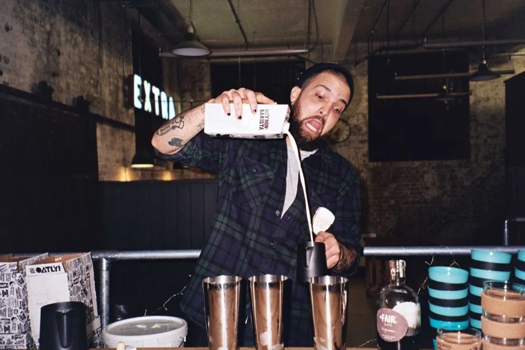 A man with tattoos is pouring cream into a glass of coffee at a cafe or bar. There are several glasses of coffee on the counter, with stacked cups and various items behind him.