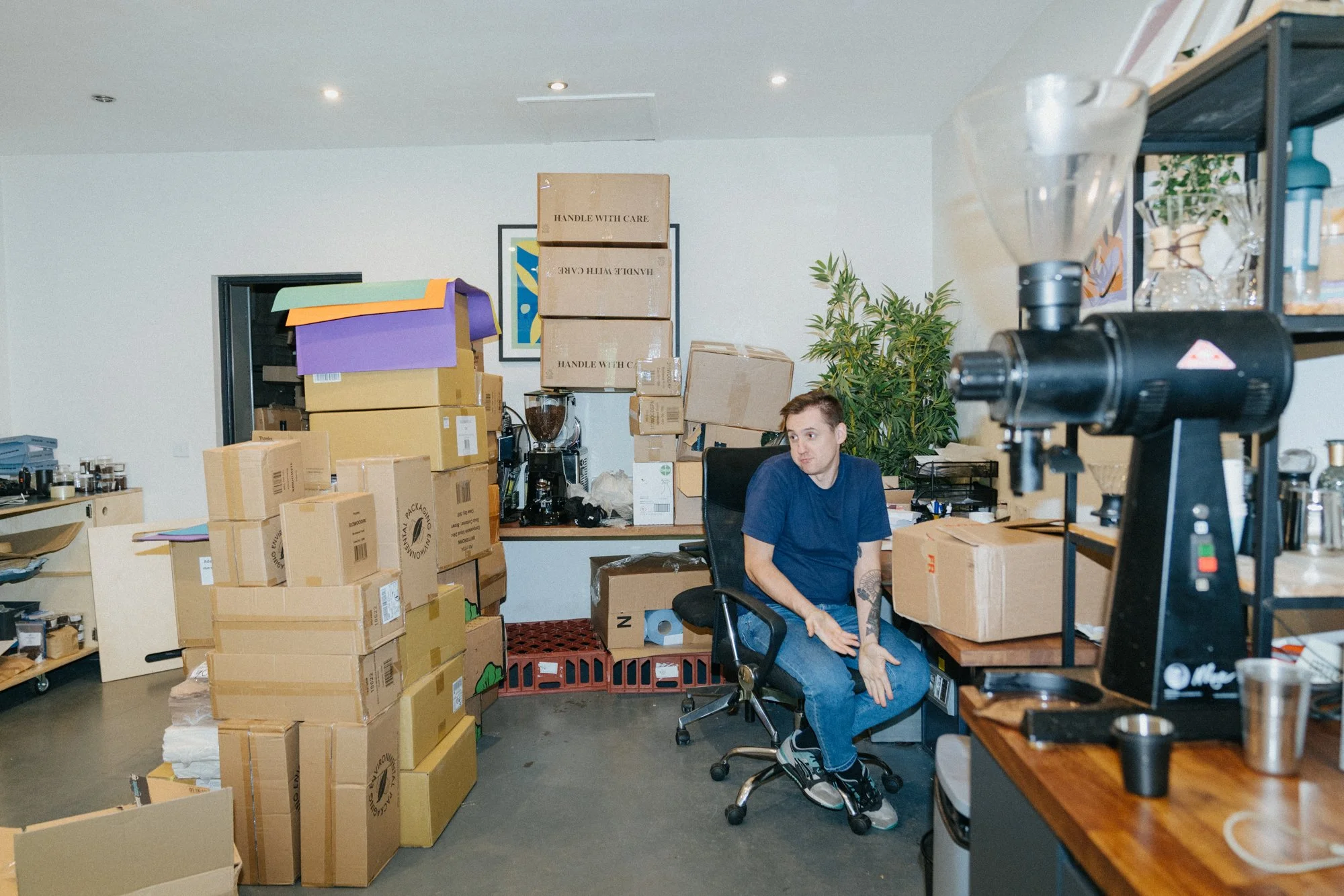 A man sitting on an office chair surrounded by stacked cardboard boxes in a cluttered workspace.