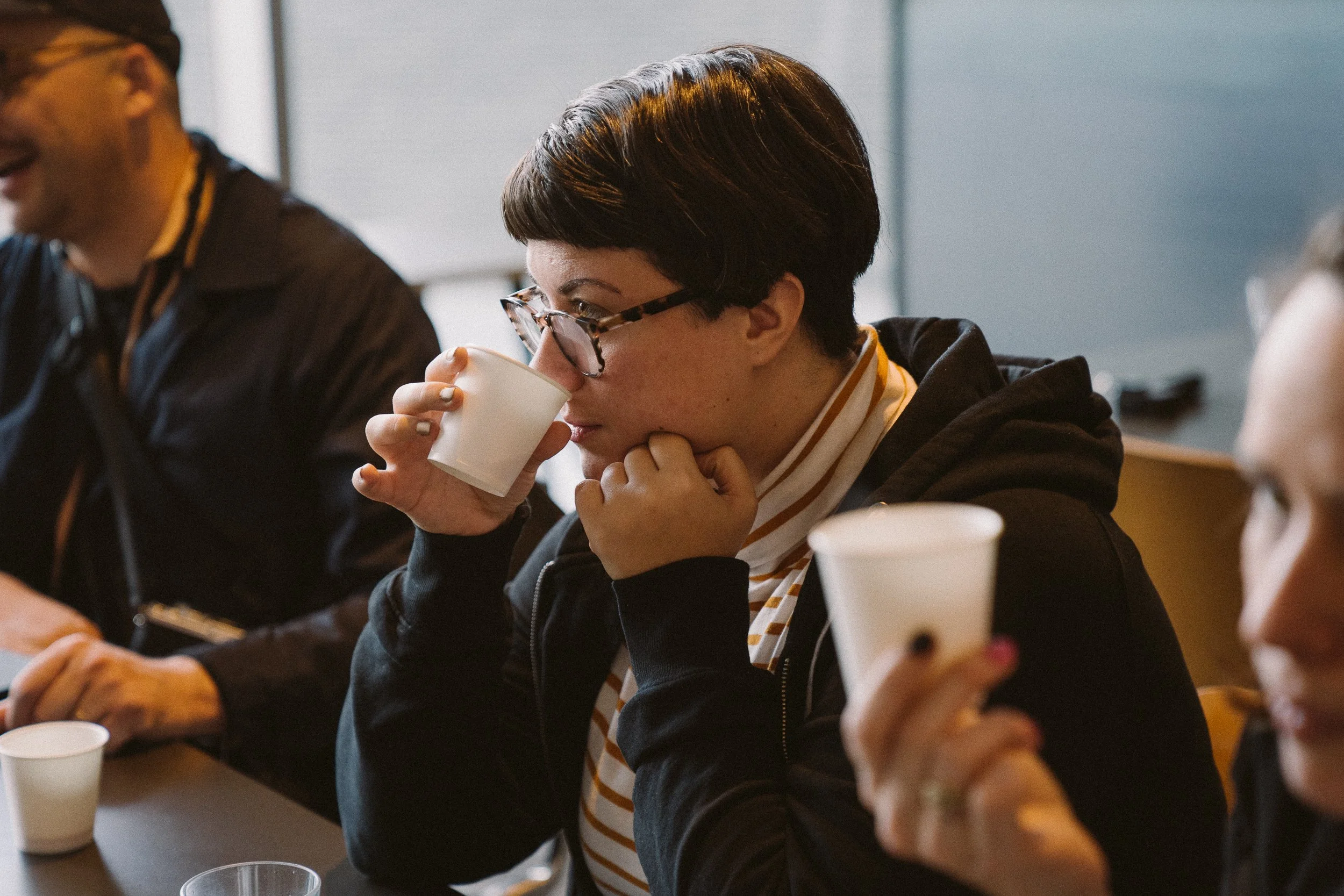 A woman with short dark hair, glasses, and a striped shirt drinks from a white paper cup during a meeting, sitting at a table with other people.