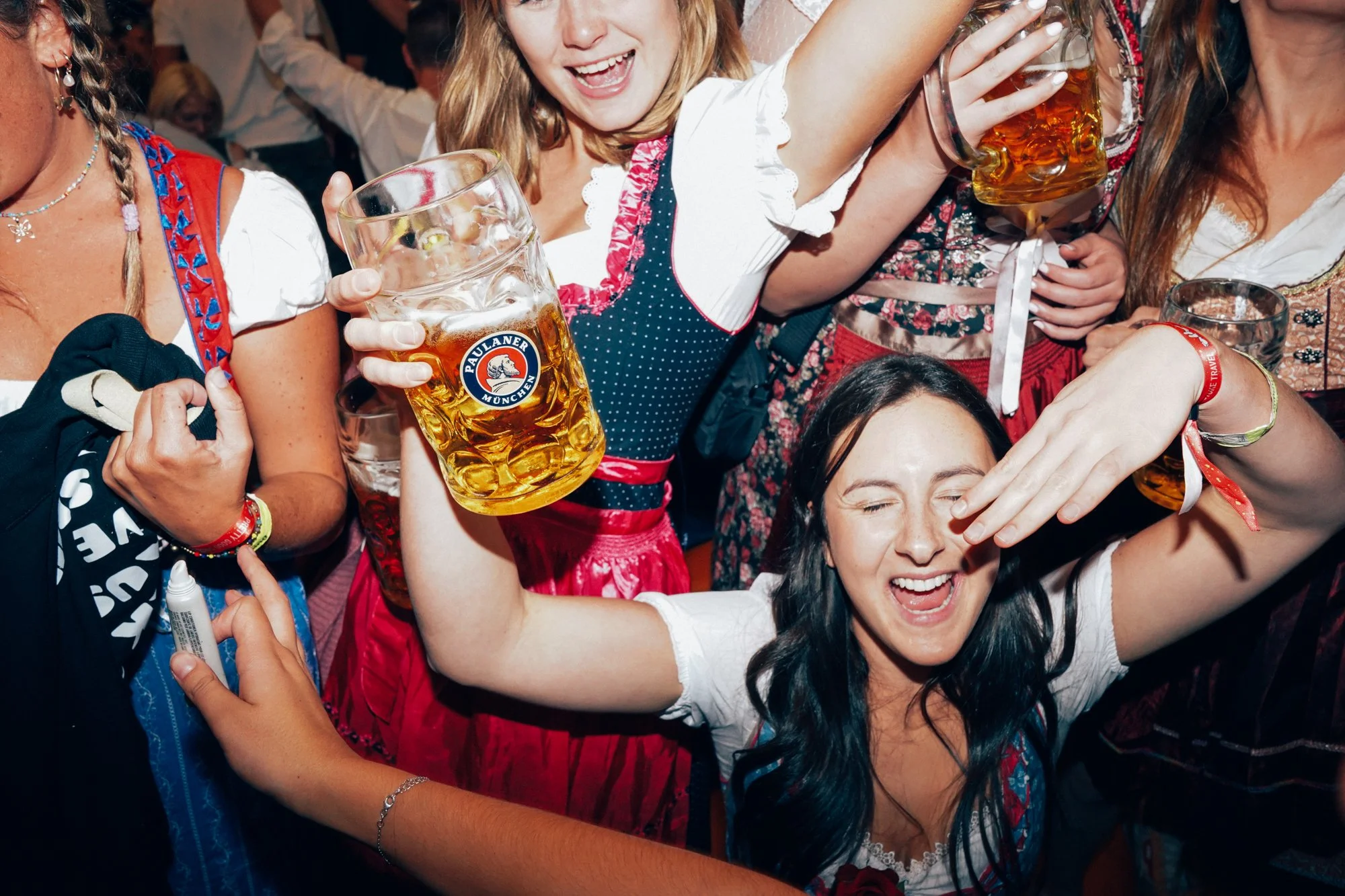 Group of women celebrating at a party wearing traditional Bavarian dresses, with some holding large mugs of beer, smiling and enjoying the moment.