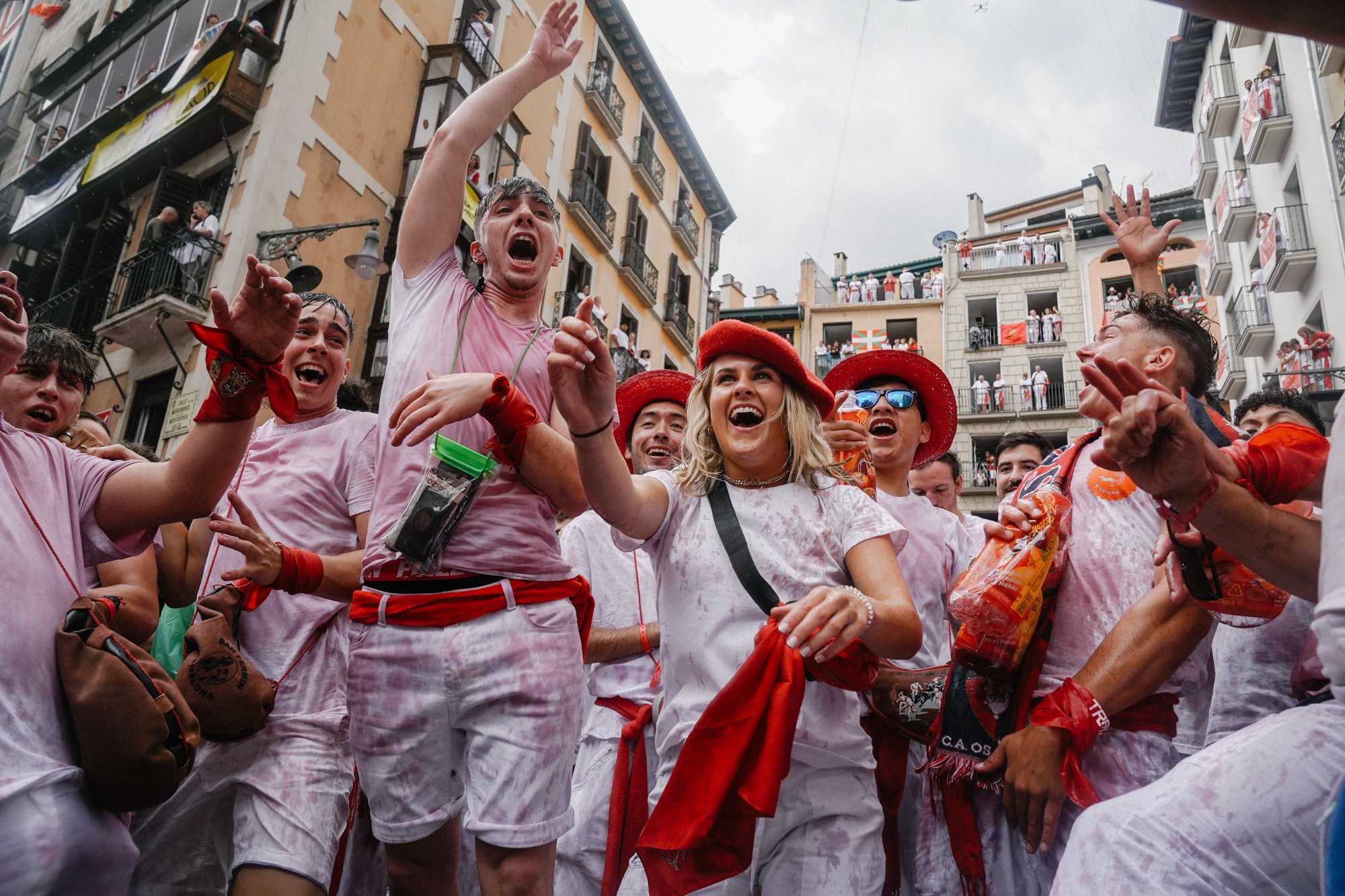 Group of people celebrating, some wearing red hats and scarves, in a lively street scene with balconies and buildings in the background, possibly during a festival or event.