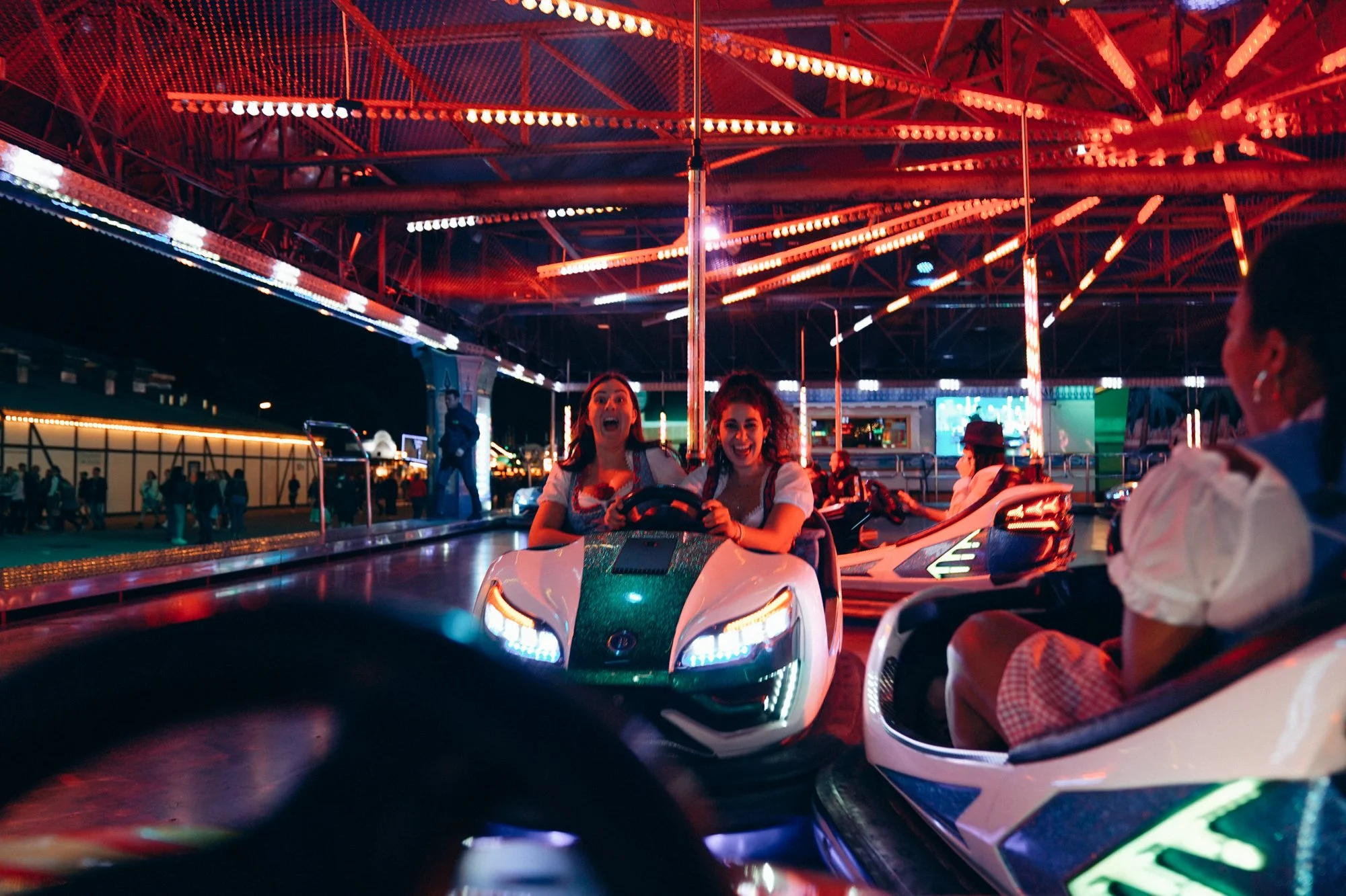 Two young women are enjoying a ride on bumper cars at a carnival or amusement park at night, with bright colorful lights and other rides visible in the background.