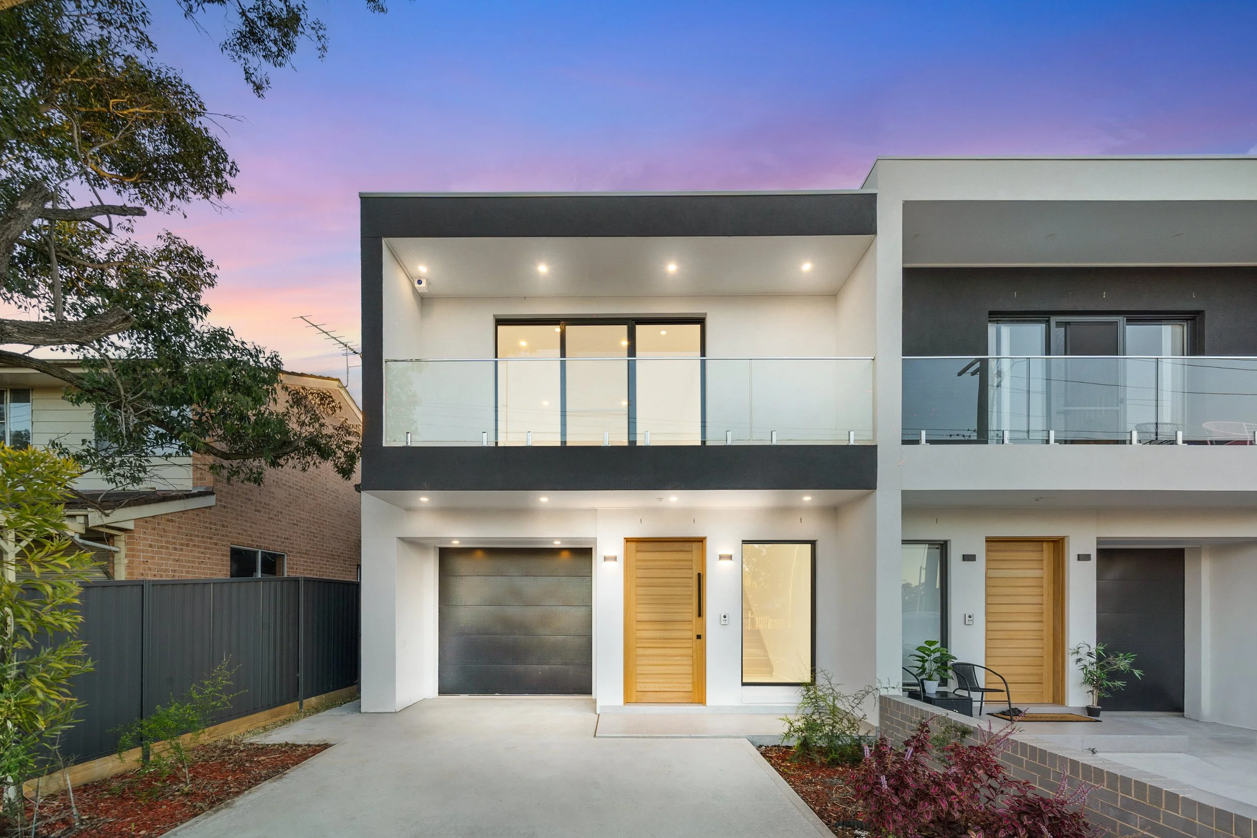 Modern two-story house with white and black exterior, wooden front doors, glass balcony, and minimal landscaping at sunset.