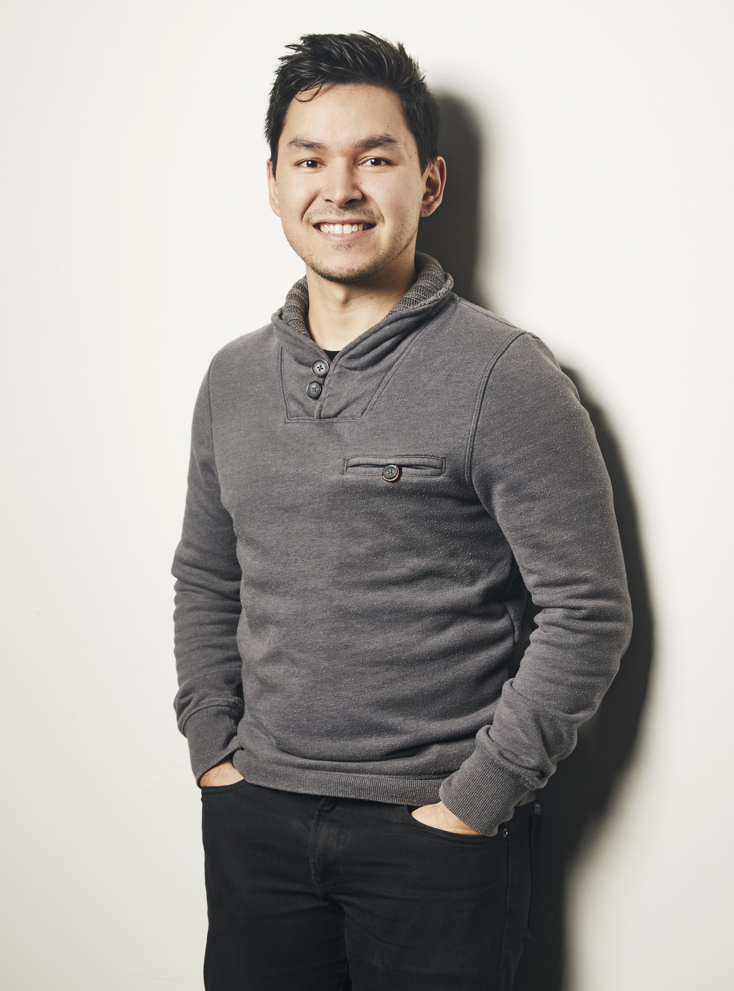 A young man with dark hair and a light beard, smiling, wearing a gray pullover with a partially buttoned collar, standing against a white wall with one hand in his pocket.