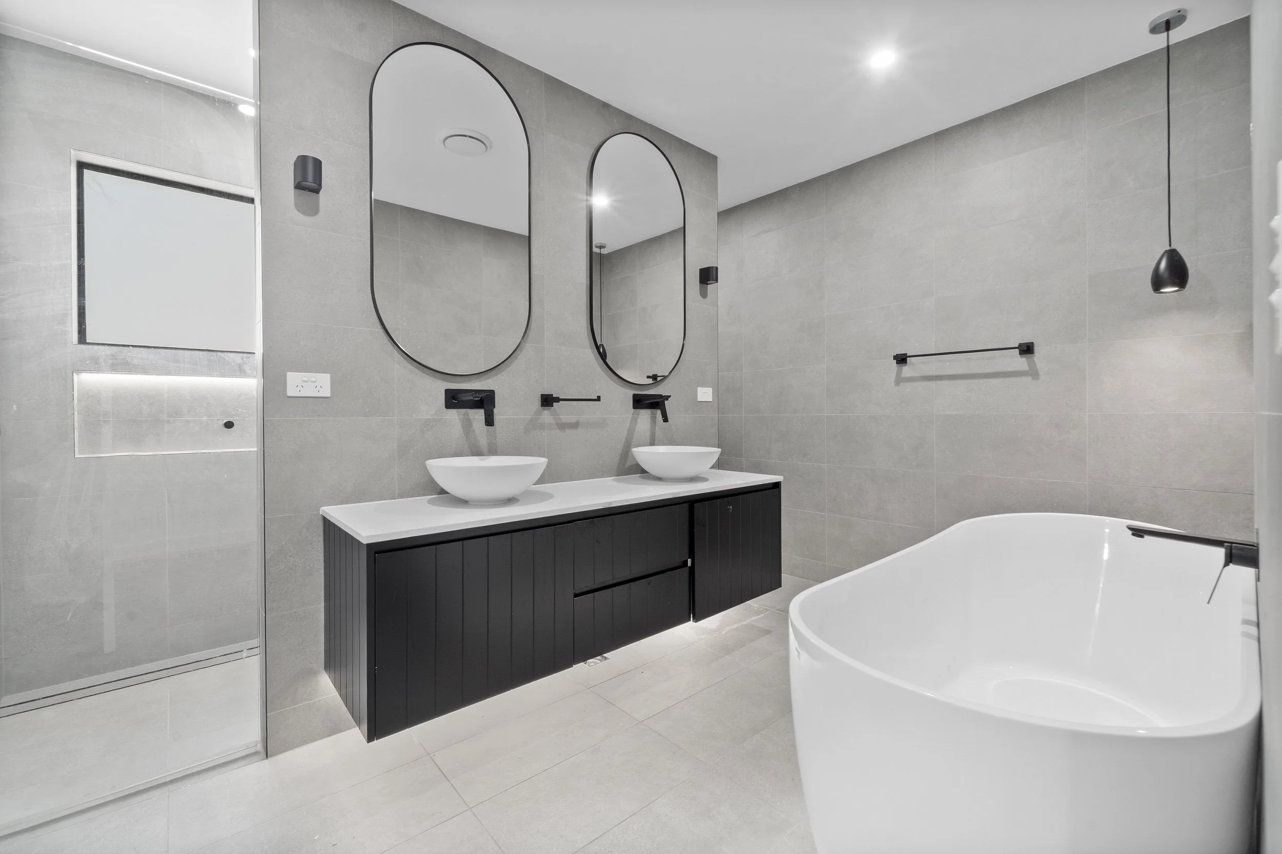 Modern bathroom with double vanity, two oval mirrors, vessel sinks, black fixtures, a bathtub, and a walk-in shower with glass door.