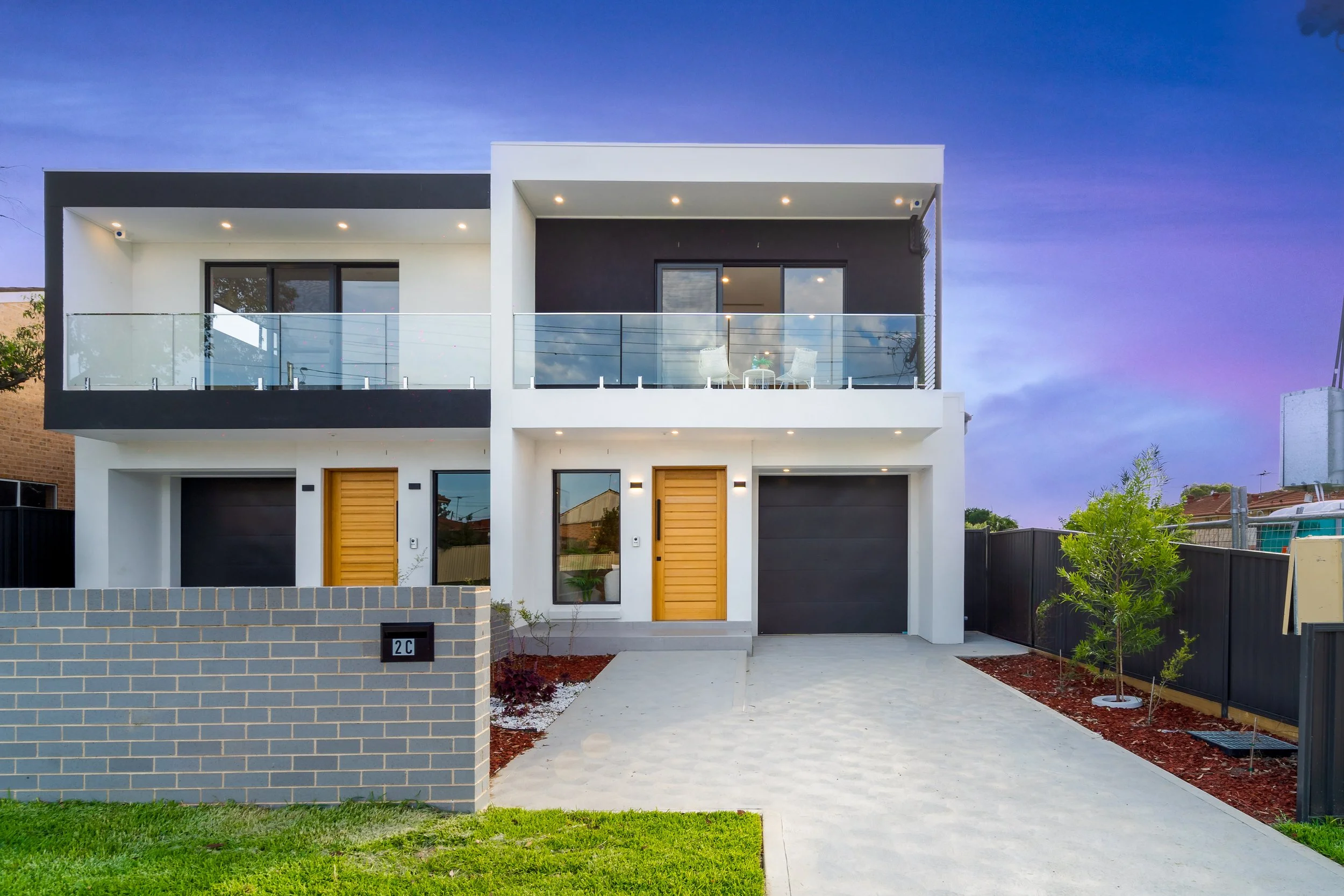 Modern two-story house with white and black exterior, wooden doors, glass balcony, and a paved driveway at dusk with a sunset sky.