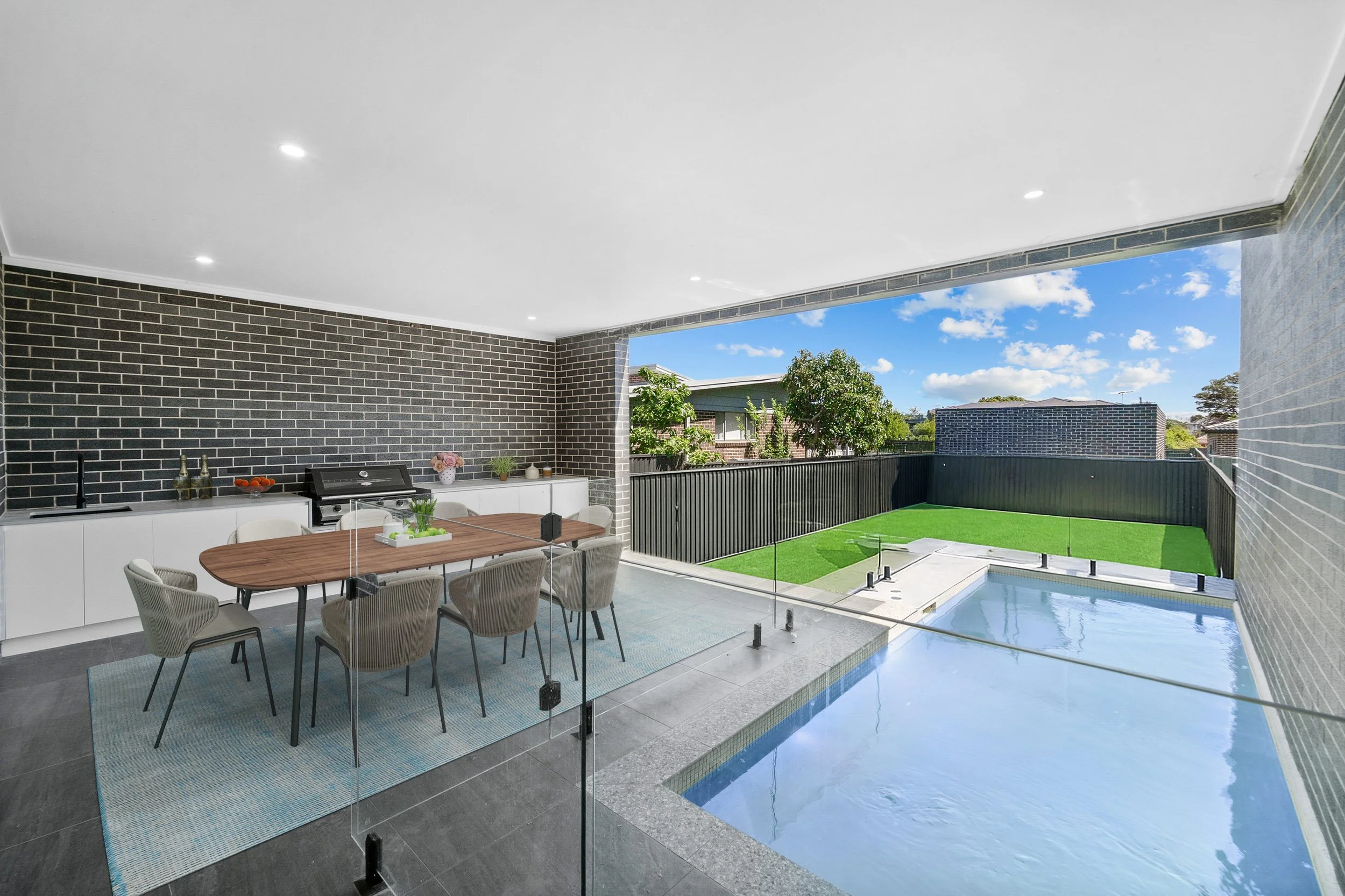 Modern outdoor patio with dining area, small pool, and fenced backyard with grass and trees under a blue sky.