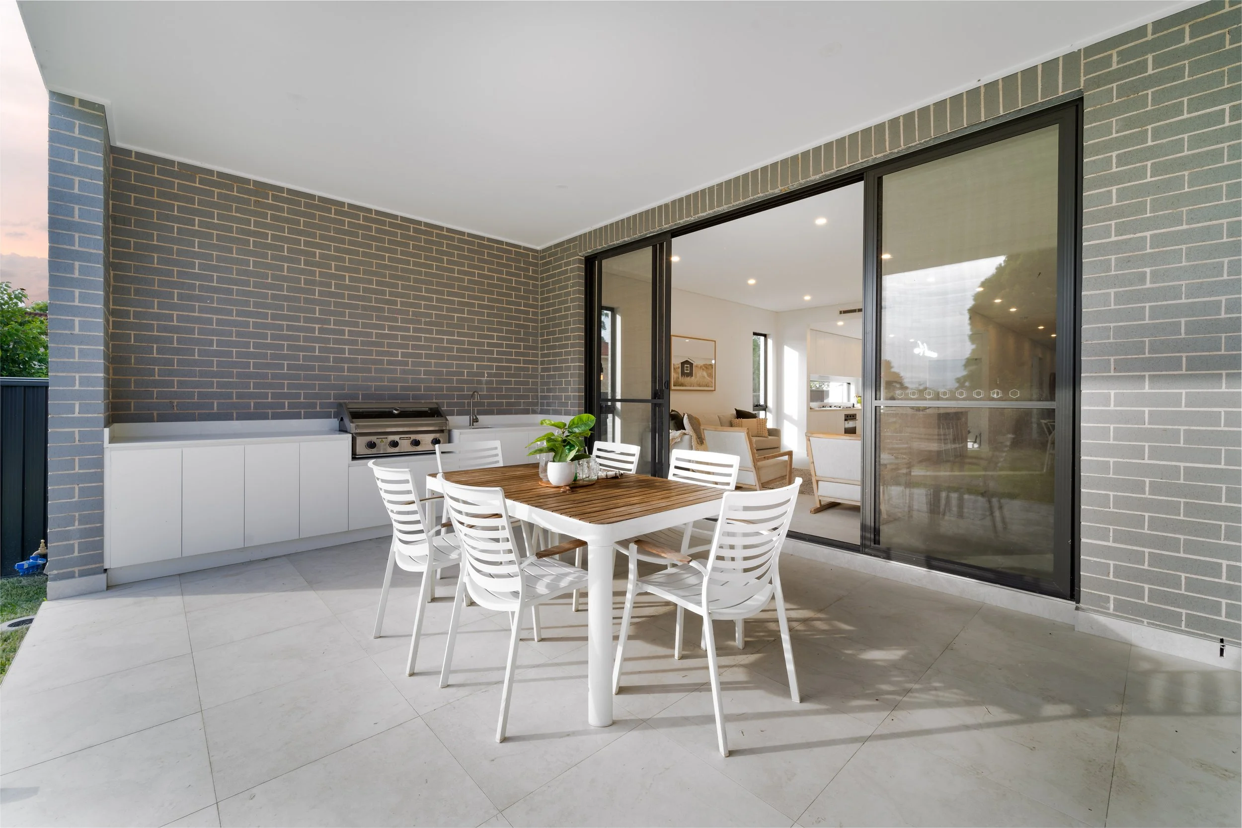 Covered outdoor patio with a white dining table, six white chairs, and a potted plant in the center. There is a built-in outdoor kitchen with a stainless steel grill and a sink, against a gray brick wall. The patio has large tiled flooring, and slidi