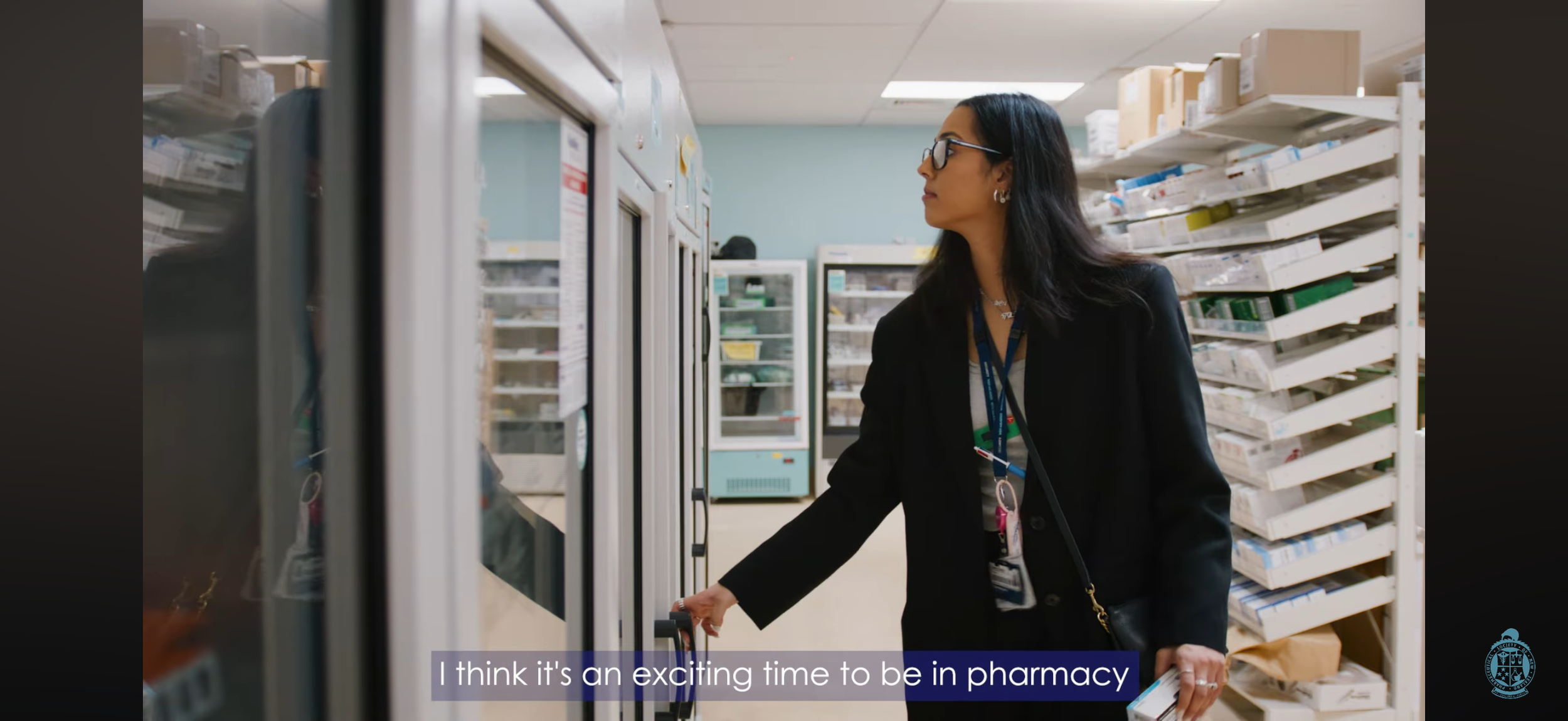 A woman in professional attire shopping in a pharmacy aisle, pushing a shopping cart, with shelves of medication and supplies in the background. She is wearing glasses and has a lanyard with ID badge.