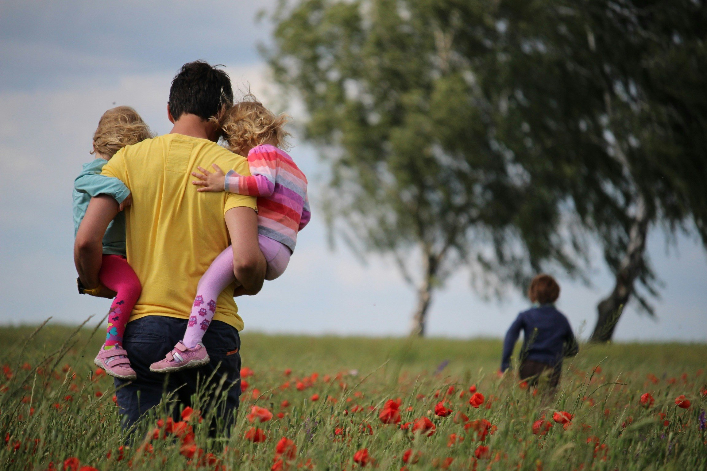 A man carrying two children on his back through a field of red flowers, with a child walking ahead and trees in the background.
