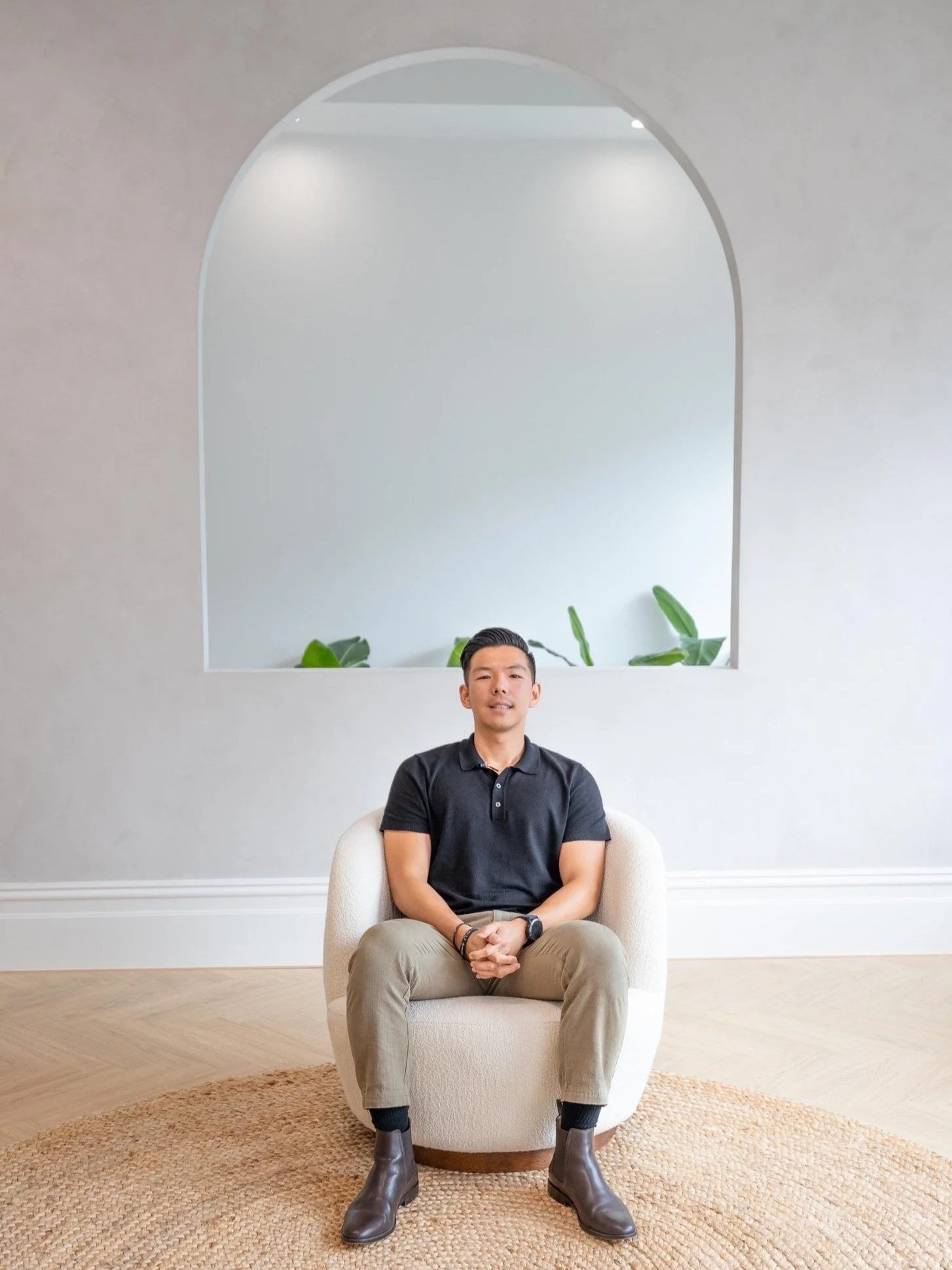 A young man sitting on a white armchair in a room with beige carpet, light grey walls, and a large arched window with plants behind him.