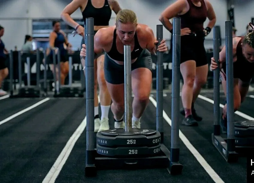 Two women in workout clothes participating in a fitness class at an indoor gym. The woman in the foreground is pushing a weighted sled, while the woman in the background runs towards her. The gym has a high ceiling with industrial lighting and a crowd of people in the background.