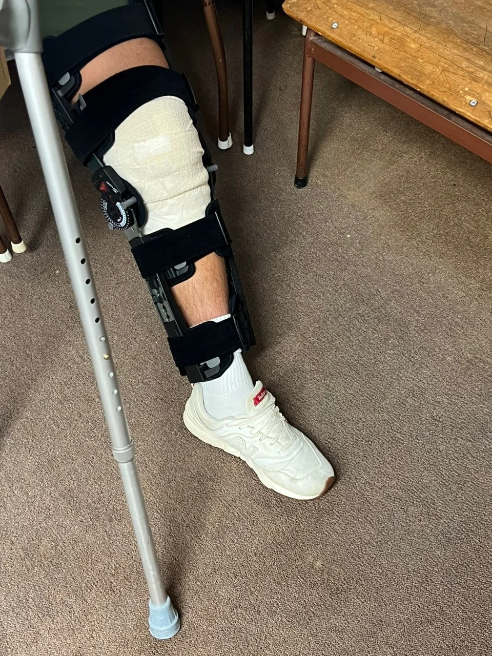A person's leg with a knee brace and crutches, wearing a white sneaker and white sock, in a room with brown carpet and wooden furniture.