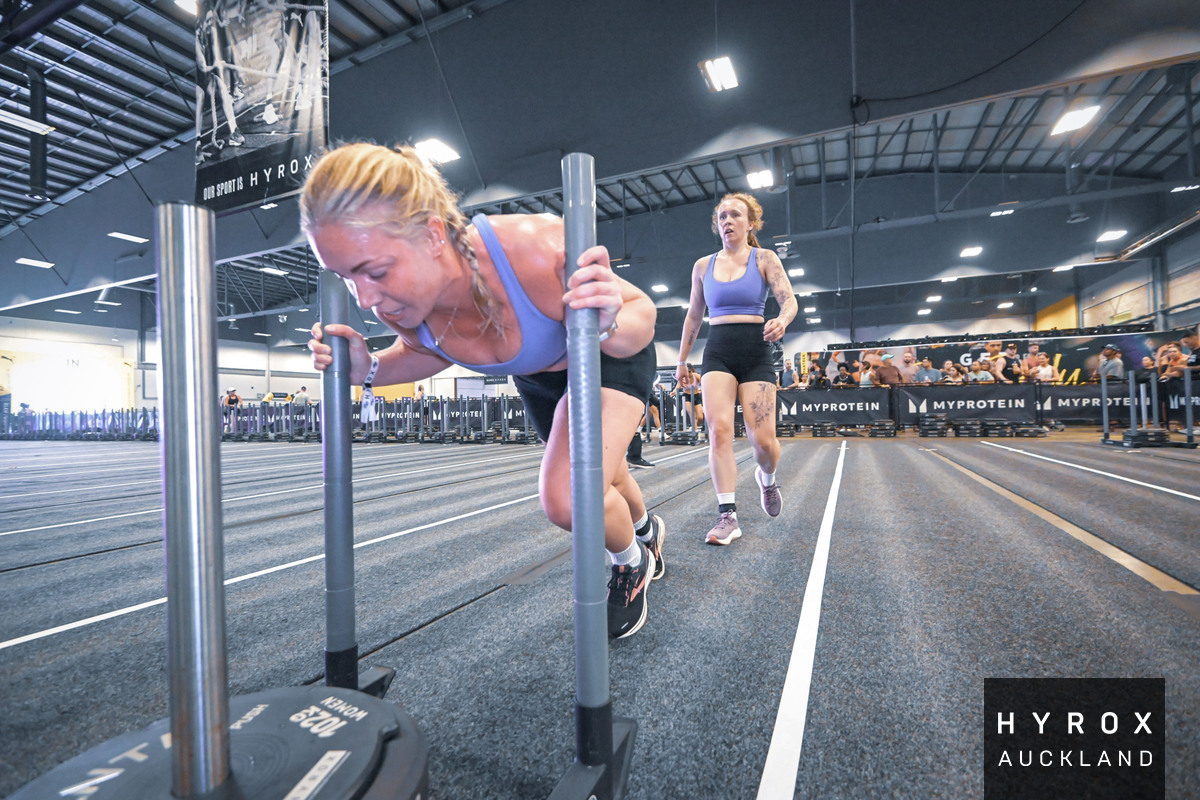 Two women working out at an indoor gym, one is pushing a weighted sled, and the other is walking behind her, both wearing athletic clothing.