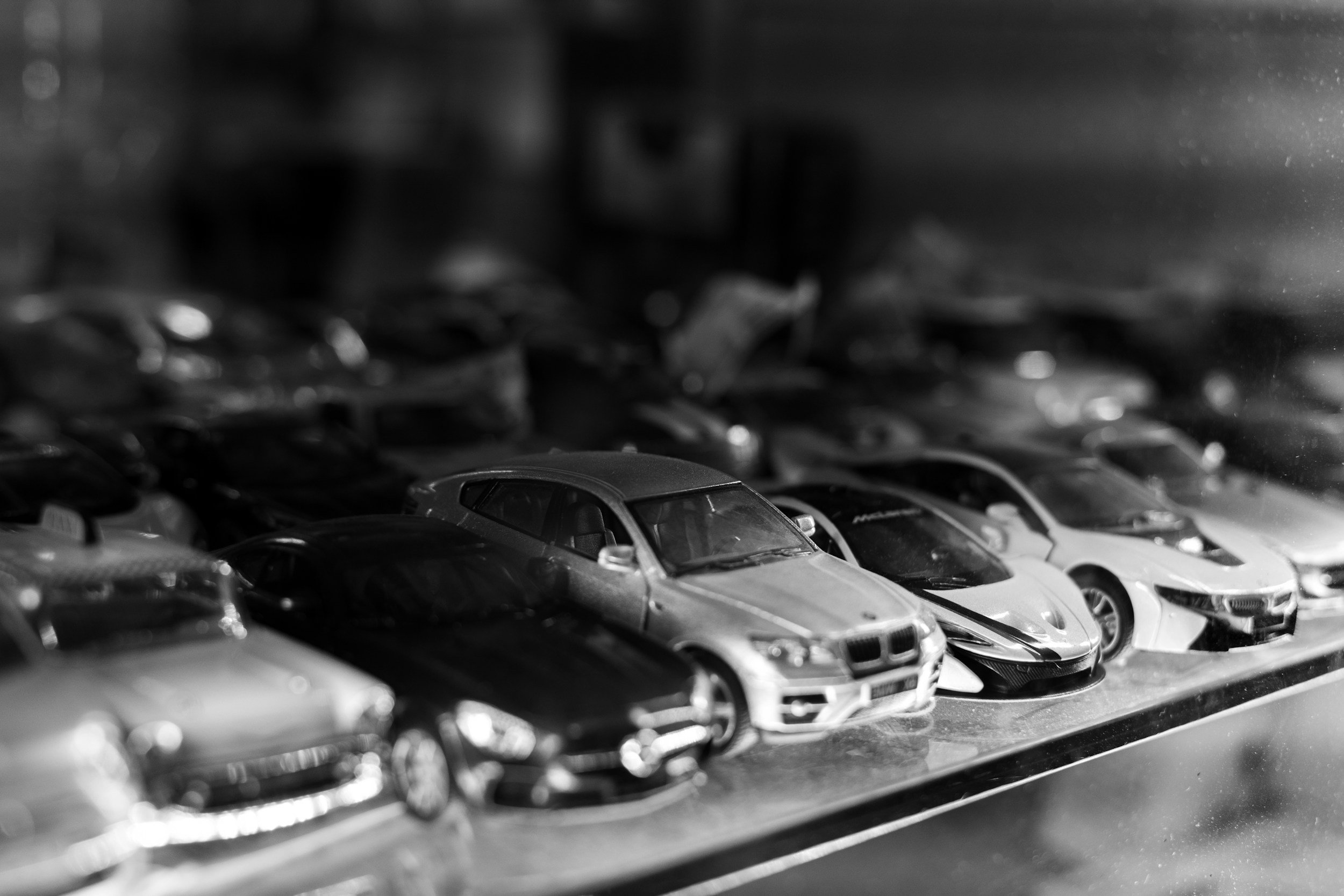Black and white photograph of a collection of toy car models on a shelf, with front cars clearly visible and others blurred in the background.
