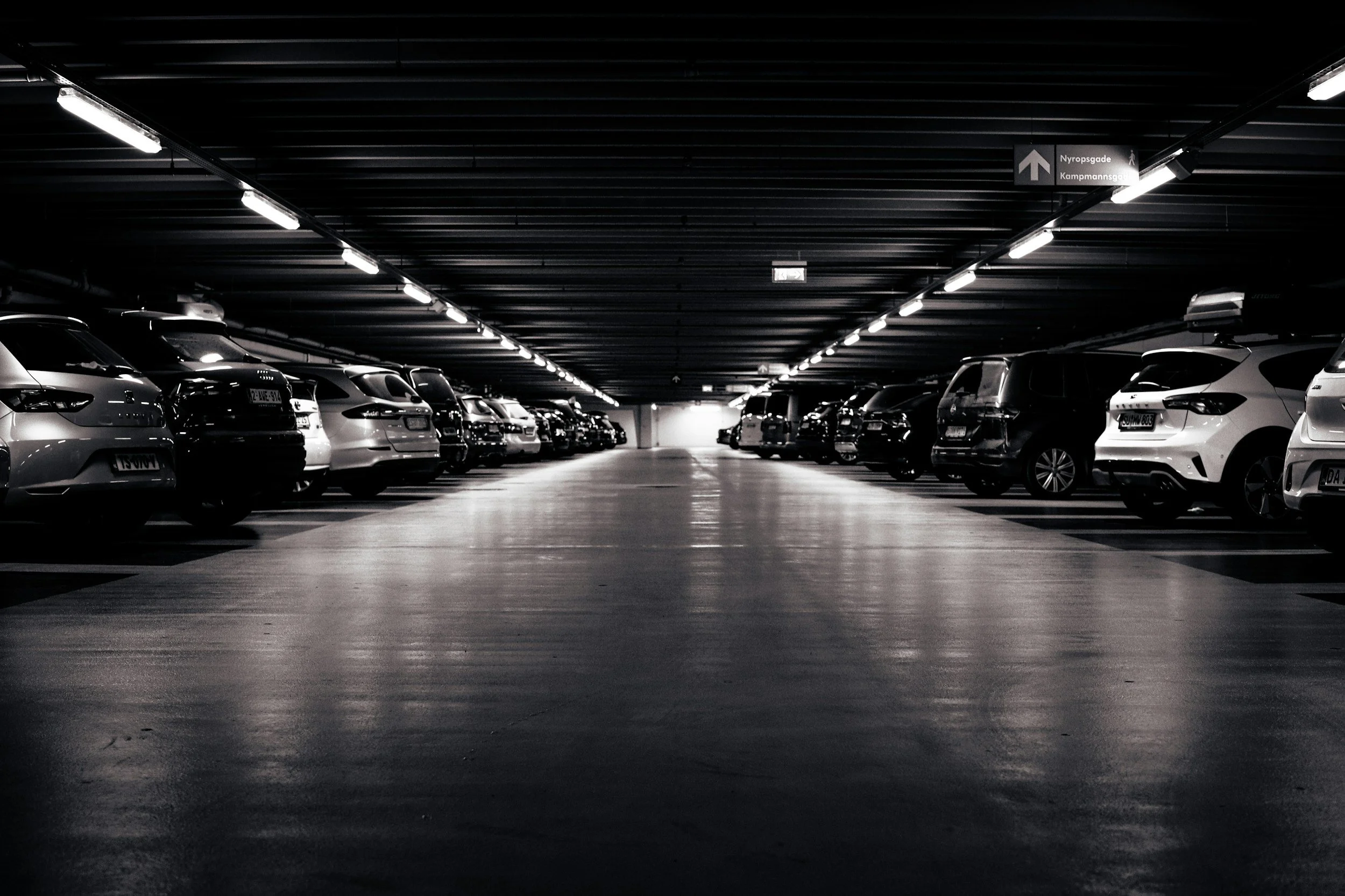 Empty indoor parking garage with cars parked along both sides, overhead lighting, and a sign directing towards Nyropsgade and Kampmannsgade.