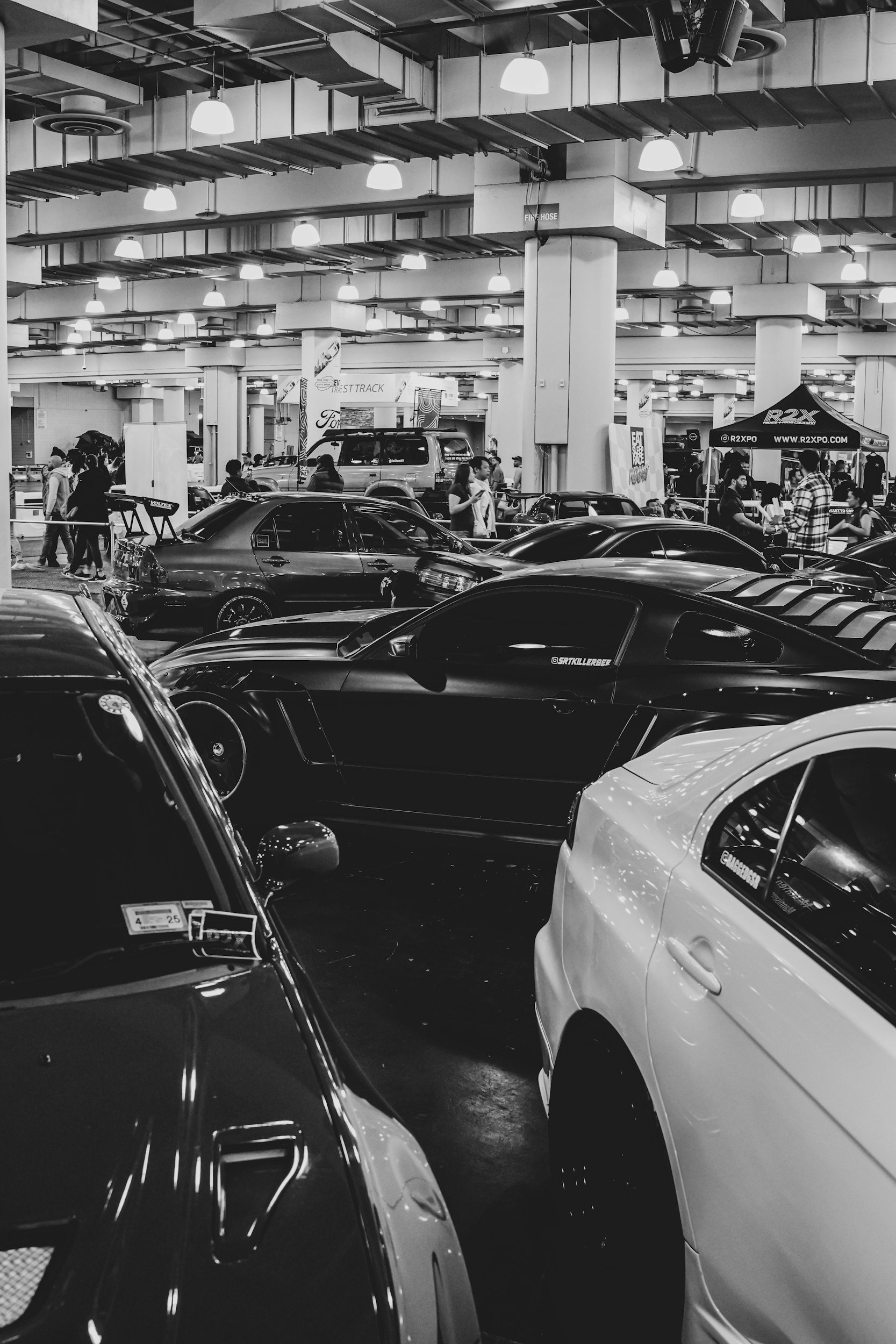 Indoor car show with various cars parked closely and people walking around, some standing near tents and booths.