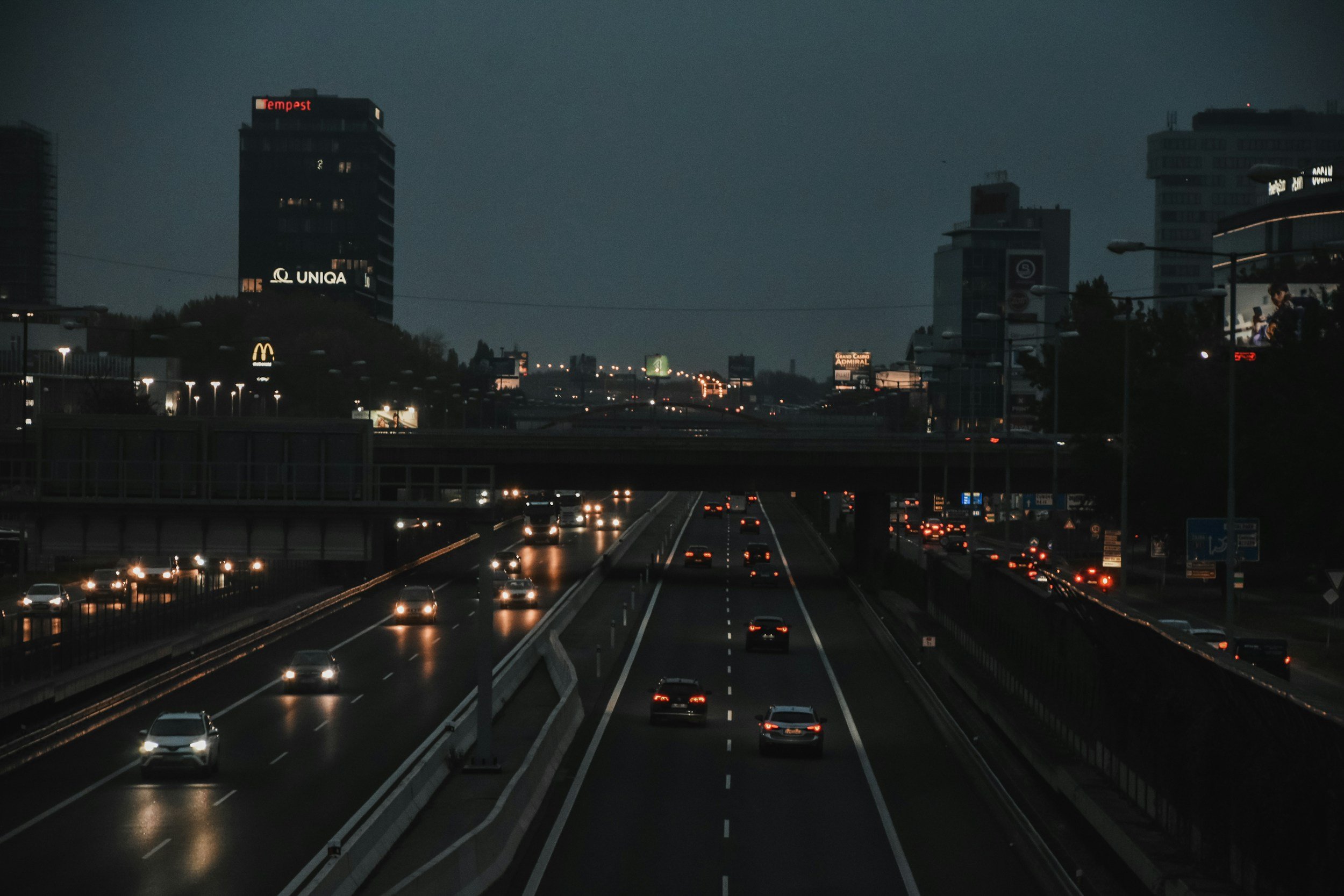 Nighttime cityscape showing a multi-lane highway with cars and city buildings in the background, some illuminated advertisements and billboards.