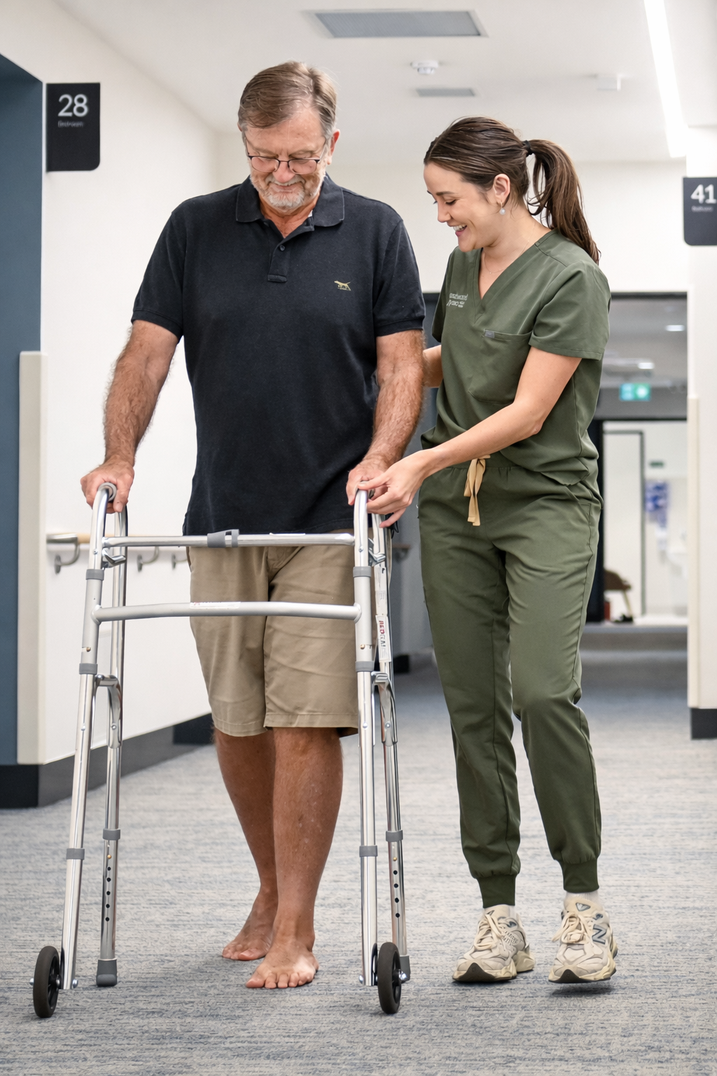 A young female healthcare worker assisting an older male patient with a walker in a hospital corridor.