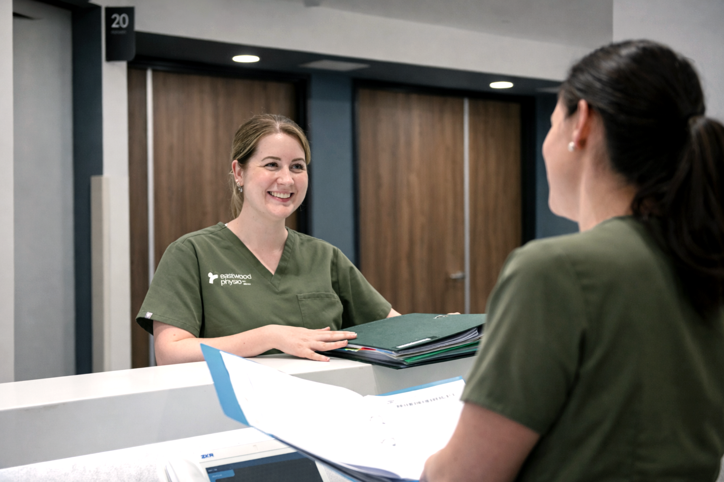A smiling woman in green scrubs sitting at a reception desk, speaking with another person in green scrubs at a medical office or clinic.