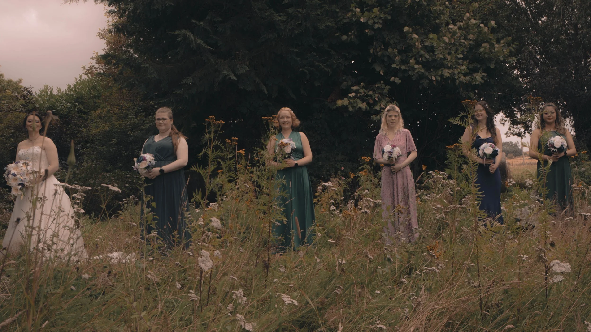 Six women dressed in formal dresses standing in a field of wildflowers with trees in the background, holding bouquets of flowers.