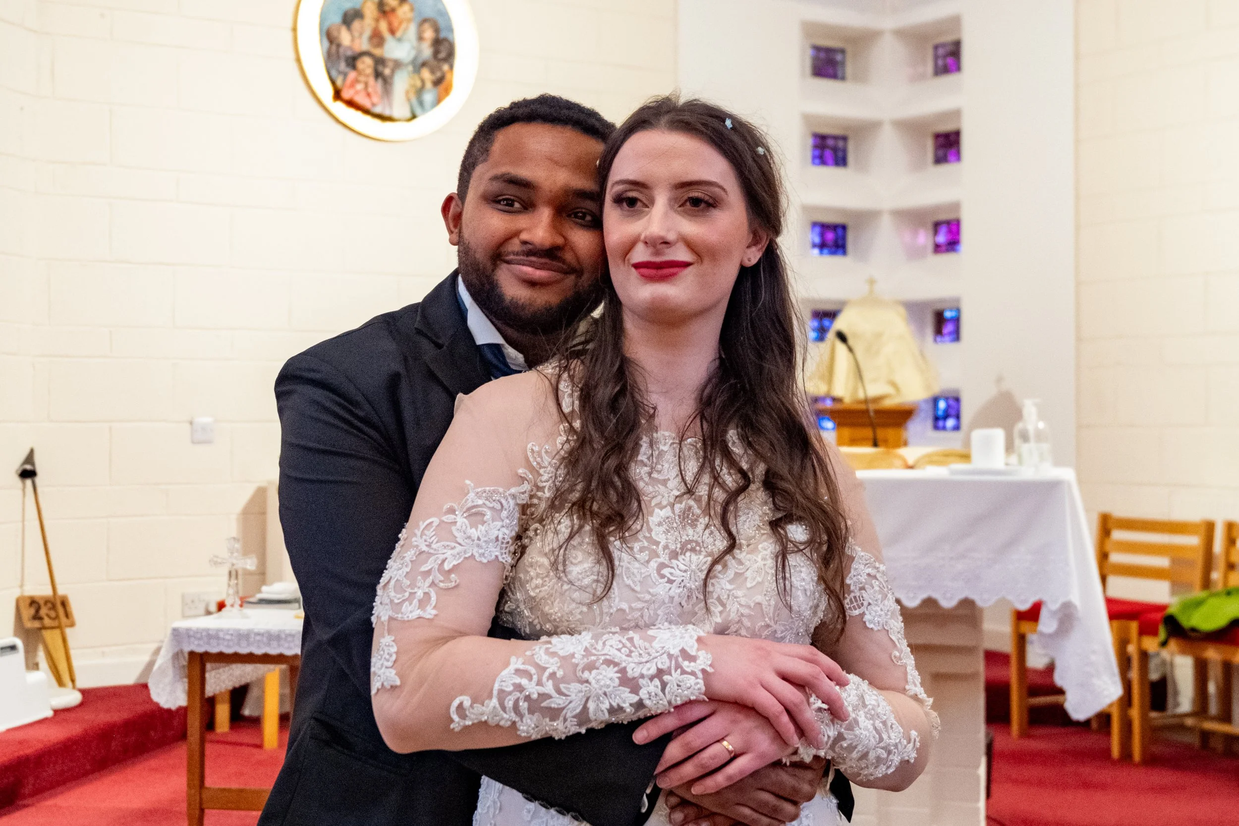 A couple standing together in a church, with the man behind the woman, both smiling and holding hands. The woman is in a white lace wedding dress, and the man is in a dark suit. The church interior is decorated with religious artwork and a table with