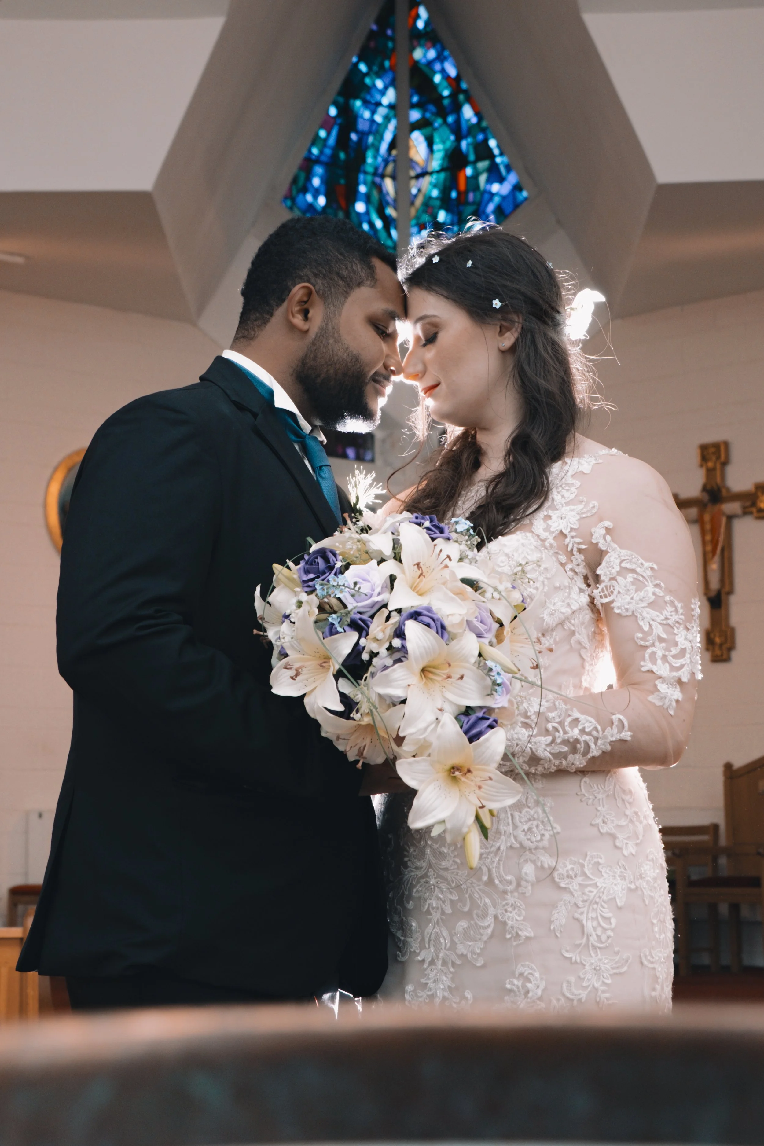 A newlywed couple standing close together inside a church, with their foreheads touching. The groom is wearing a black suit and blue tie, and the bride is wearing a white lace wedding dress, holding a bouquet of white and purple flowers. A stained gl