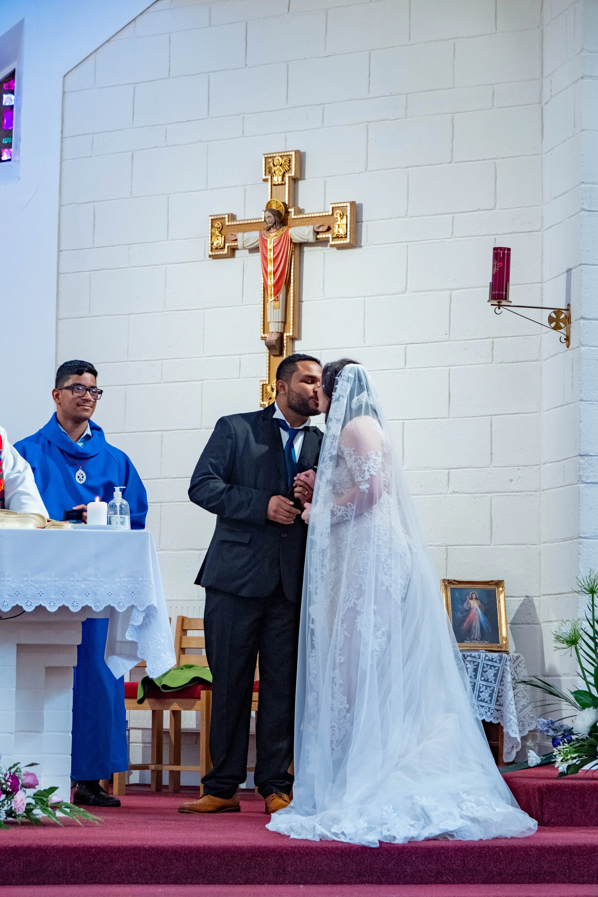 A wedding ceremony with a bride and groom kissing in a church with crucifix on the wall behind them. An altar with religious items is visible to the left, and a priest or officiant stands nearby. Flowers decorate the church interior.