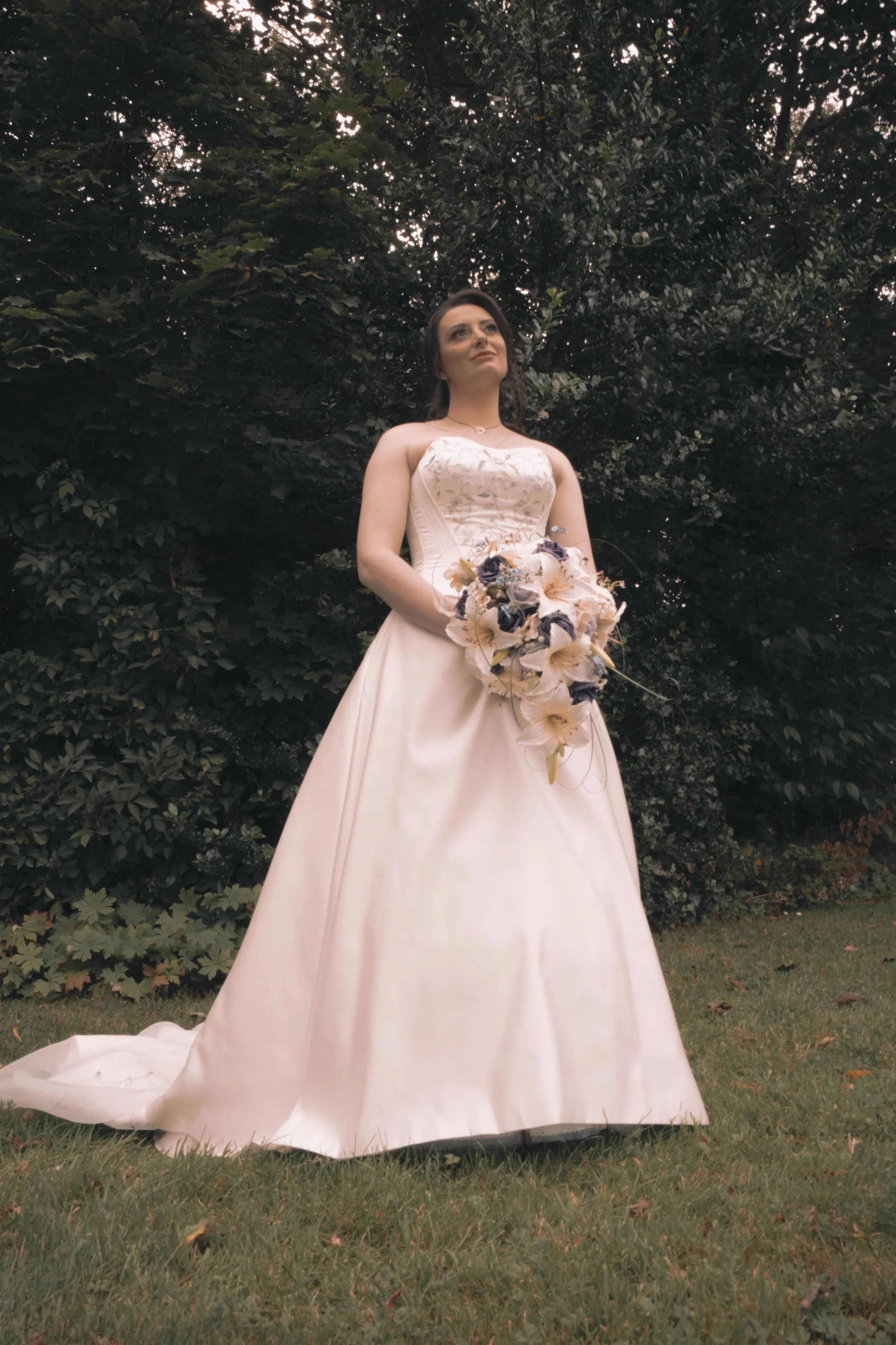 A bride in a strapless white wedding gown holding a bouquet of flowers, standing outdoors on grass with trees in the background.