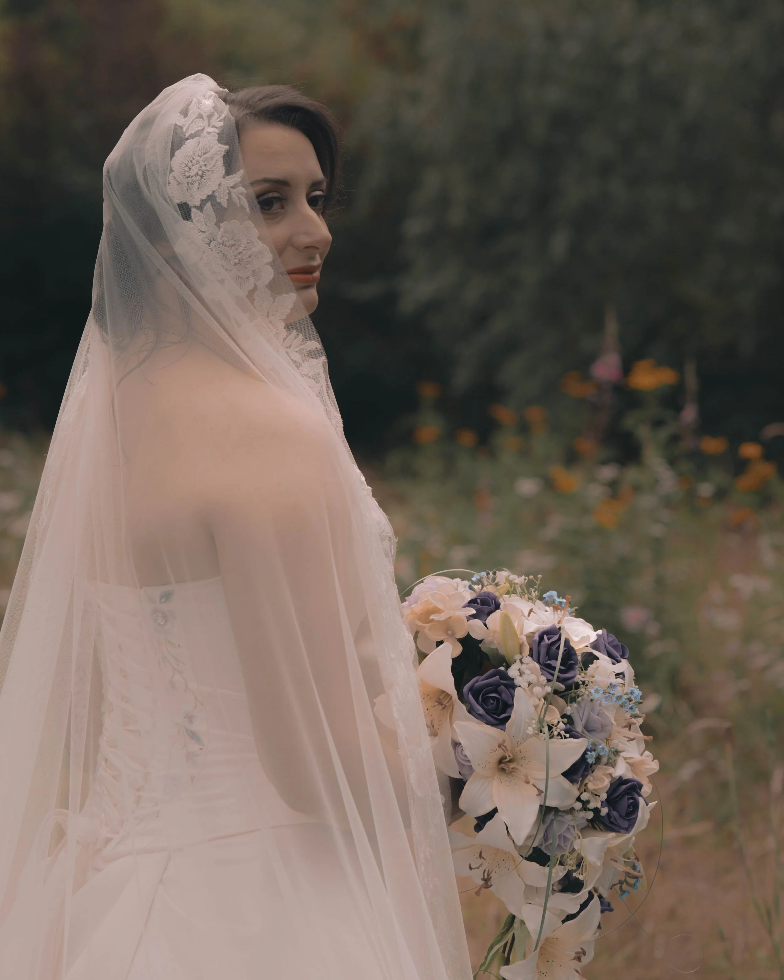 A bride with a veil and lace detailing holding a bouquet of white and purple flowers outdoors.