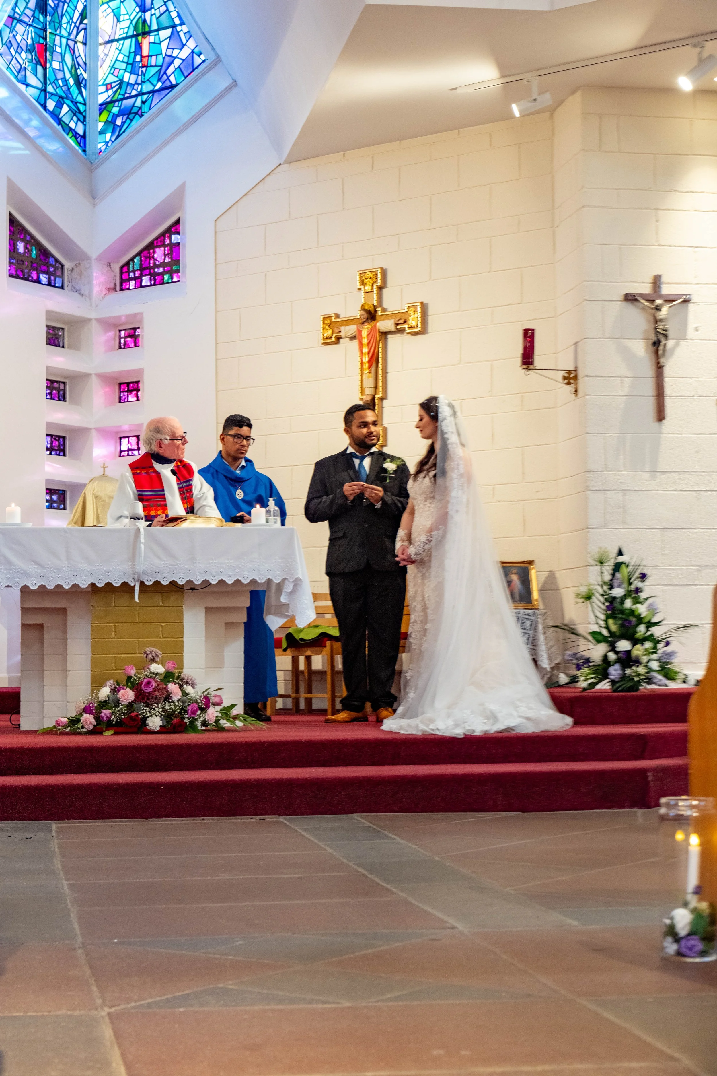 A wedding ceremony inside a church with two officiants and a couple exchanging vows. The bride wears a white wedding dress with a veil, and the groom wears a black suit. The church features stained glass windows and religious symbols, including a cro