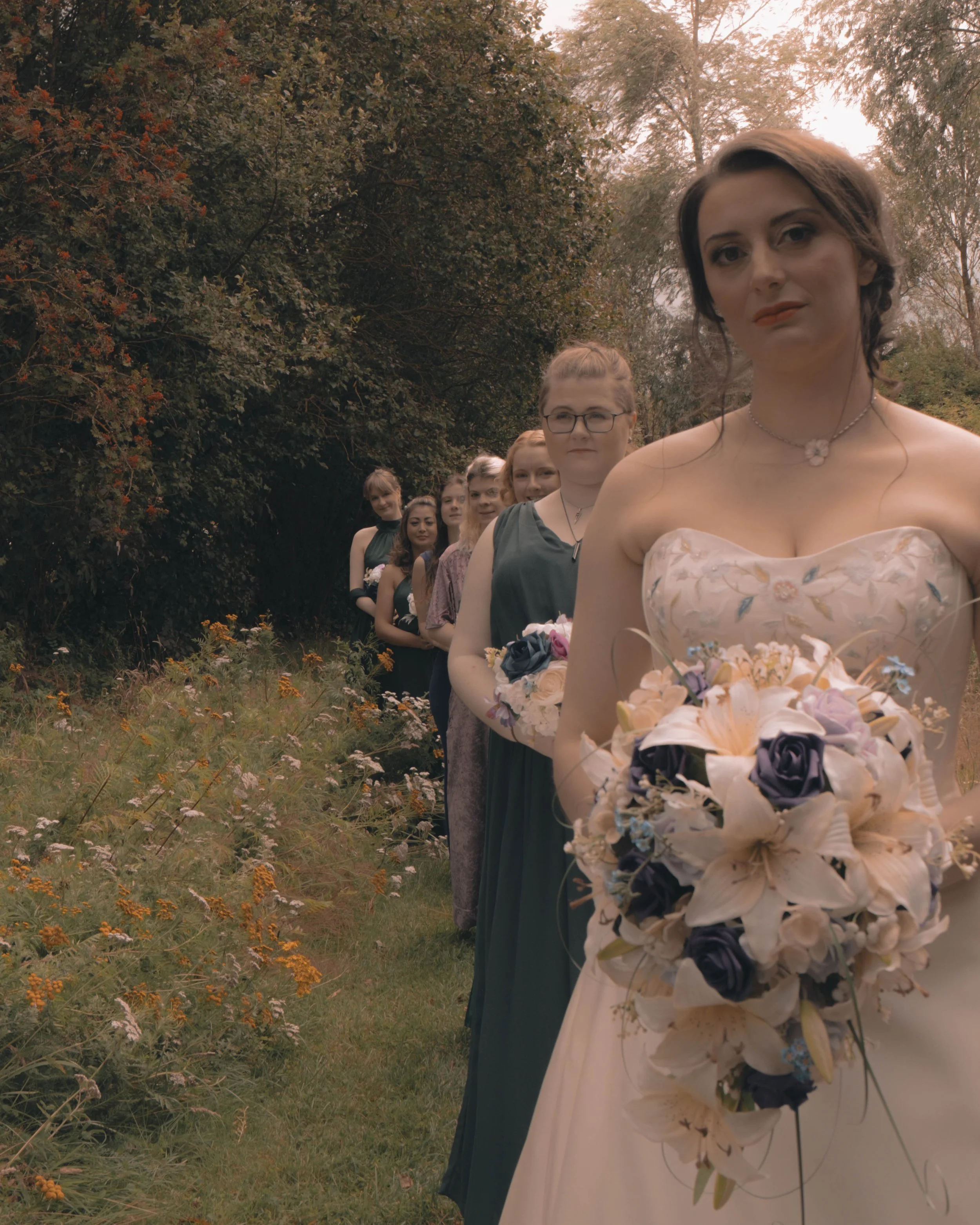 A long line of women, including a bride in a wedding dress holding a bouquet, walking outdoors in a natural setting with trees and grass.