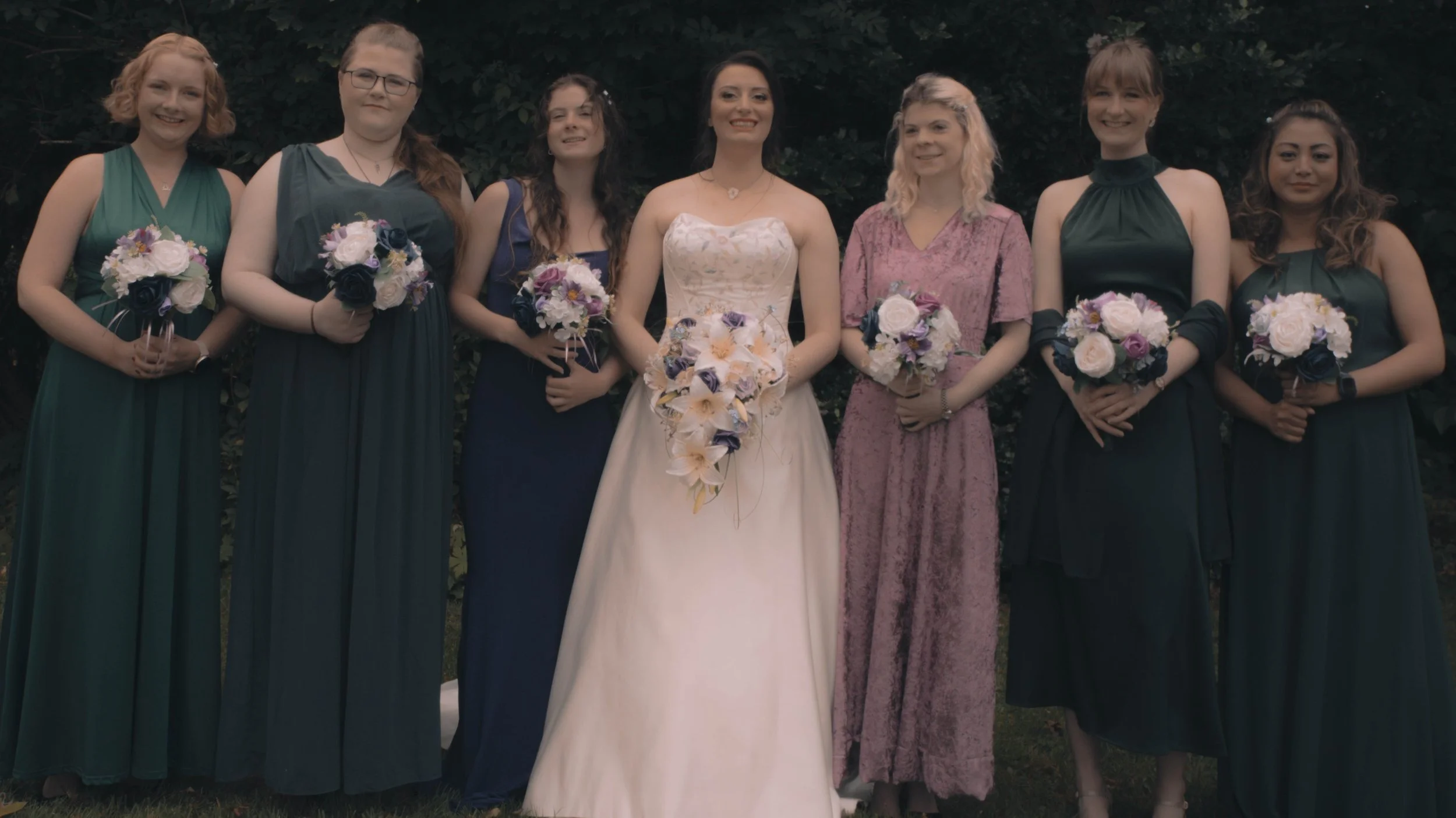 A bride and six bridesmaids standing outdoors against dark foliage, all holding bouquets of flowers.''