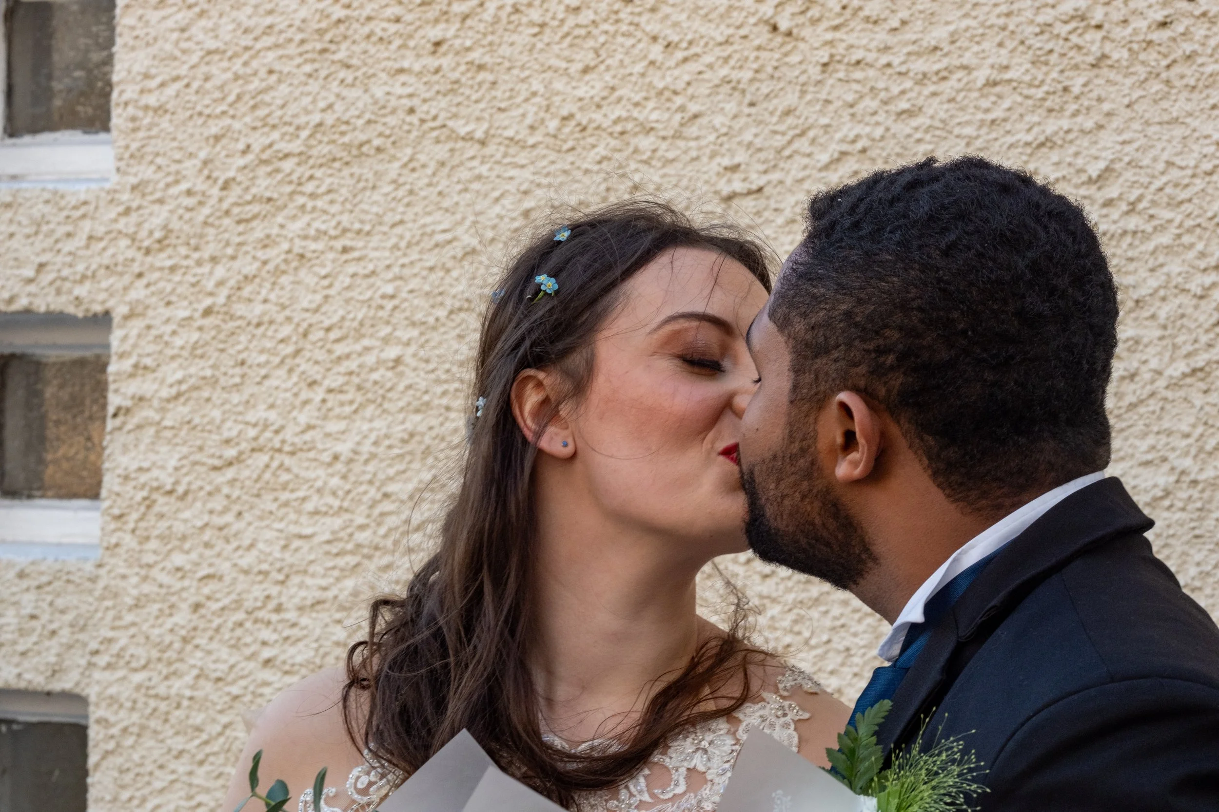 A bride and groom sharing a kiss outside, with the bride holding a bouquet, against a textured beige wall.