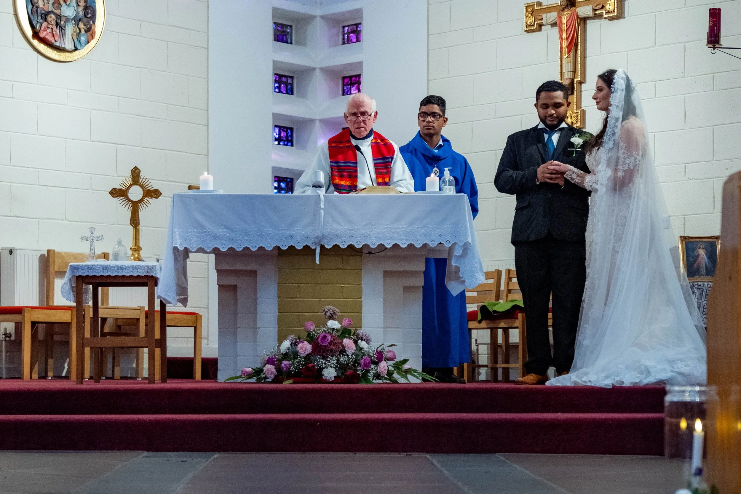 A bride and groom standing at the altar during a wedding ceremony in a church, with a priest and an altar server present.