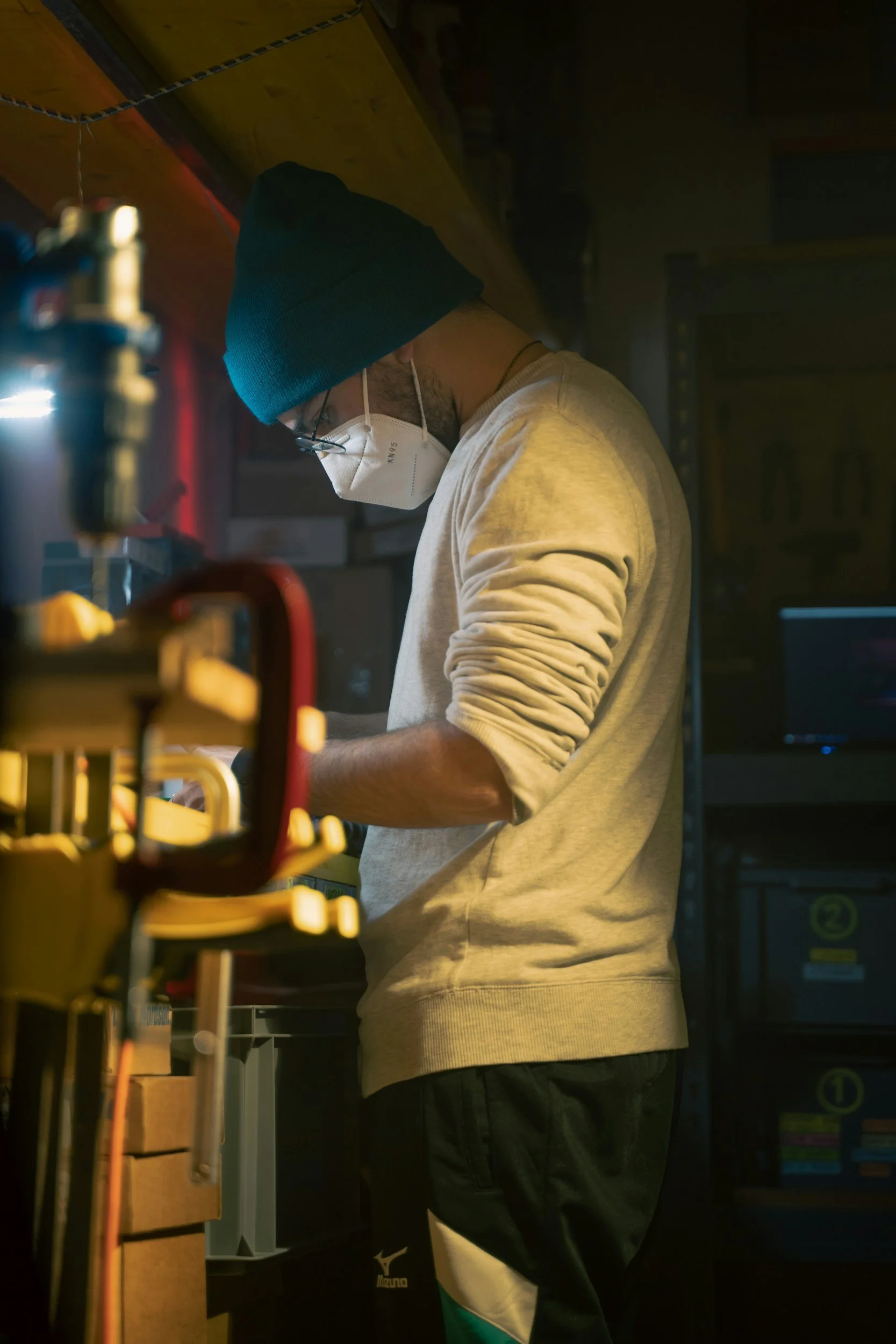 A person wearing a blue beanie, glasses, and a white face mask working in a dimly lit workshop or laboratory with yellow and red equipment around them.