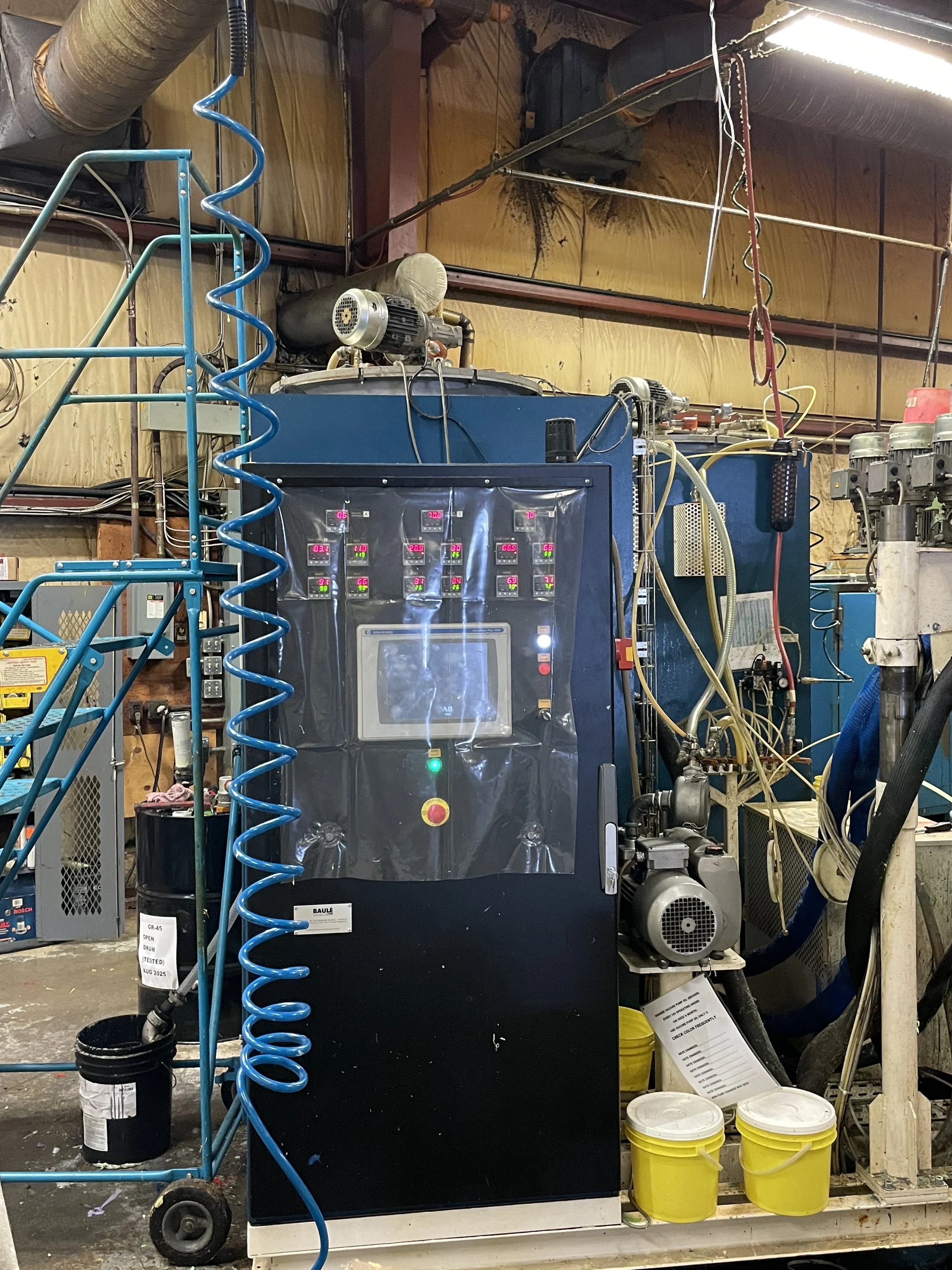 A large industrial machine with a control panel, various wires, and motors in a workshop setting. Blue coiled air hoses are hanging nearby, and yellow buckets are on the floor.